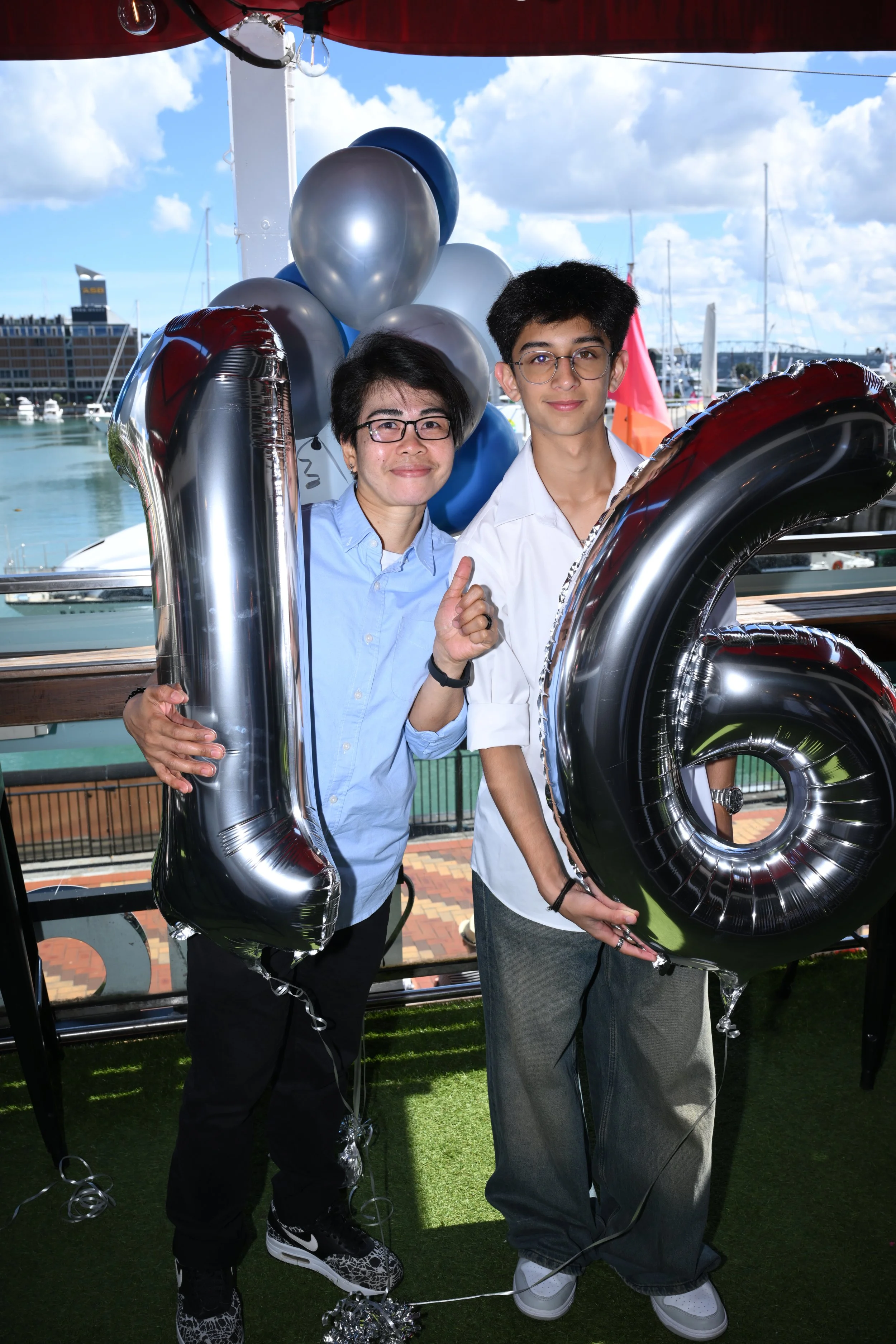 Two people holding large black and silver balloons that form the number 16, standing outdoors near a harbor with boats, a building, and a cloudy sky in the background.