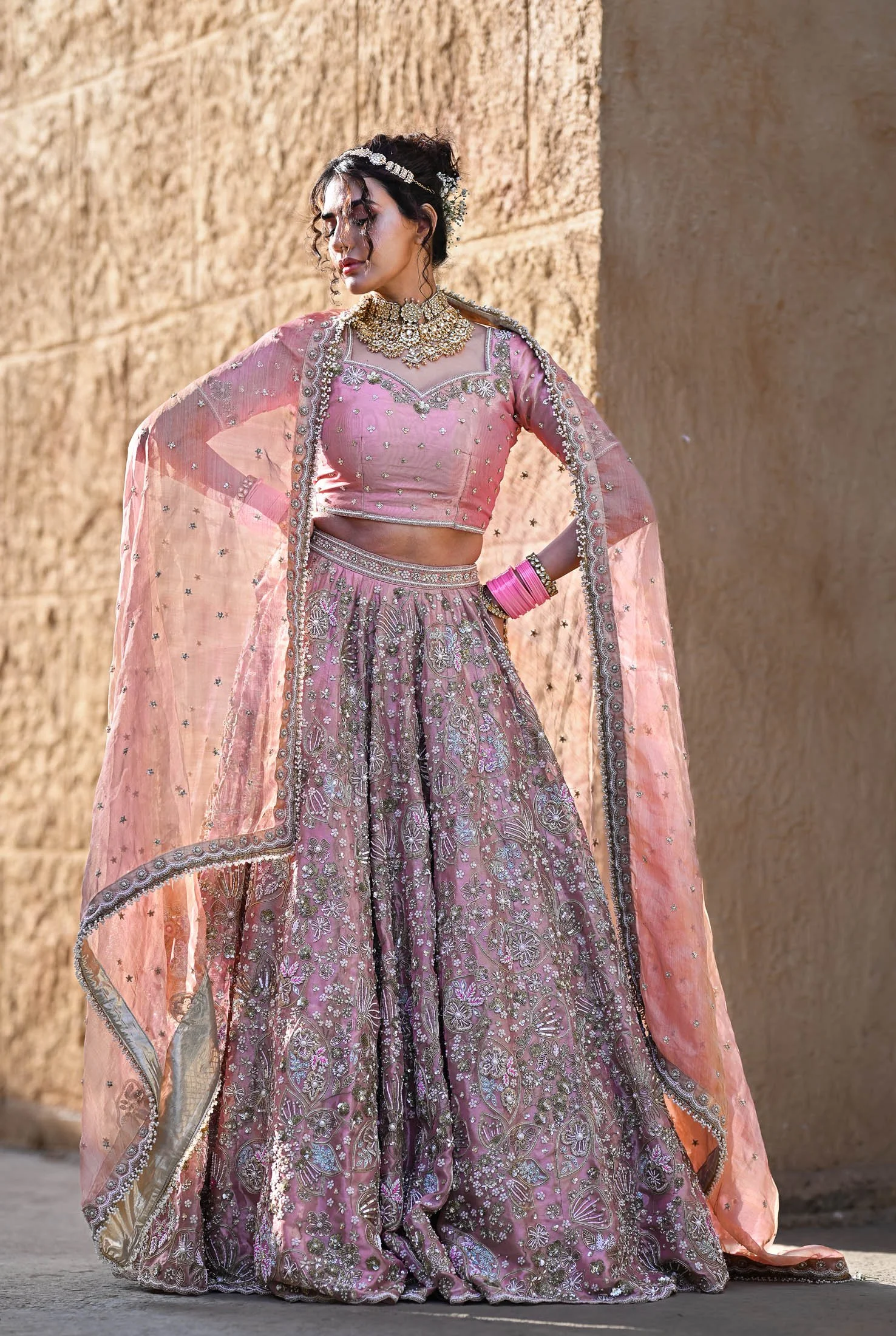 A woman wearing a traditional pink and gold embroidered Indian bridal outfit, including a lehenga, choli, dupatta, jewelry, and pink bangles, standing against a beige stone wall.