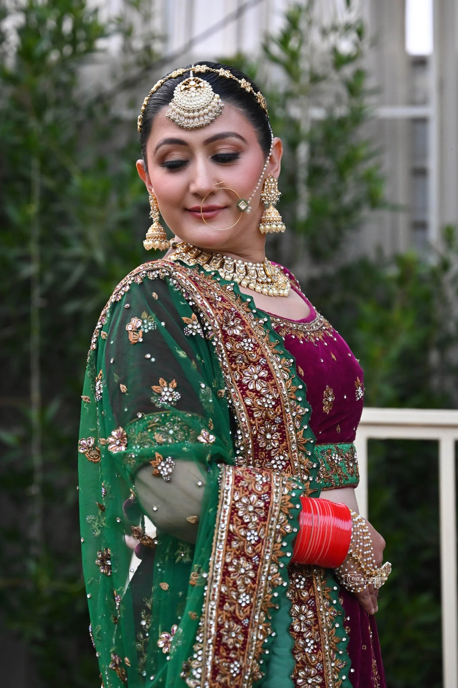 A woman dressed in traditional Indian attire, wearing a sleeveless maroon and green embroidered outfit with jewelry, including a nose ring, earrings, necklace, and bangles, standing outdoors with greenery in the background.