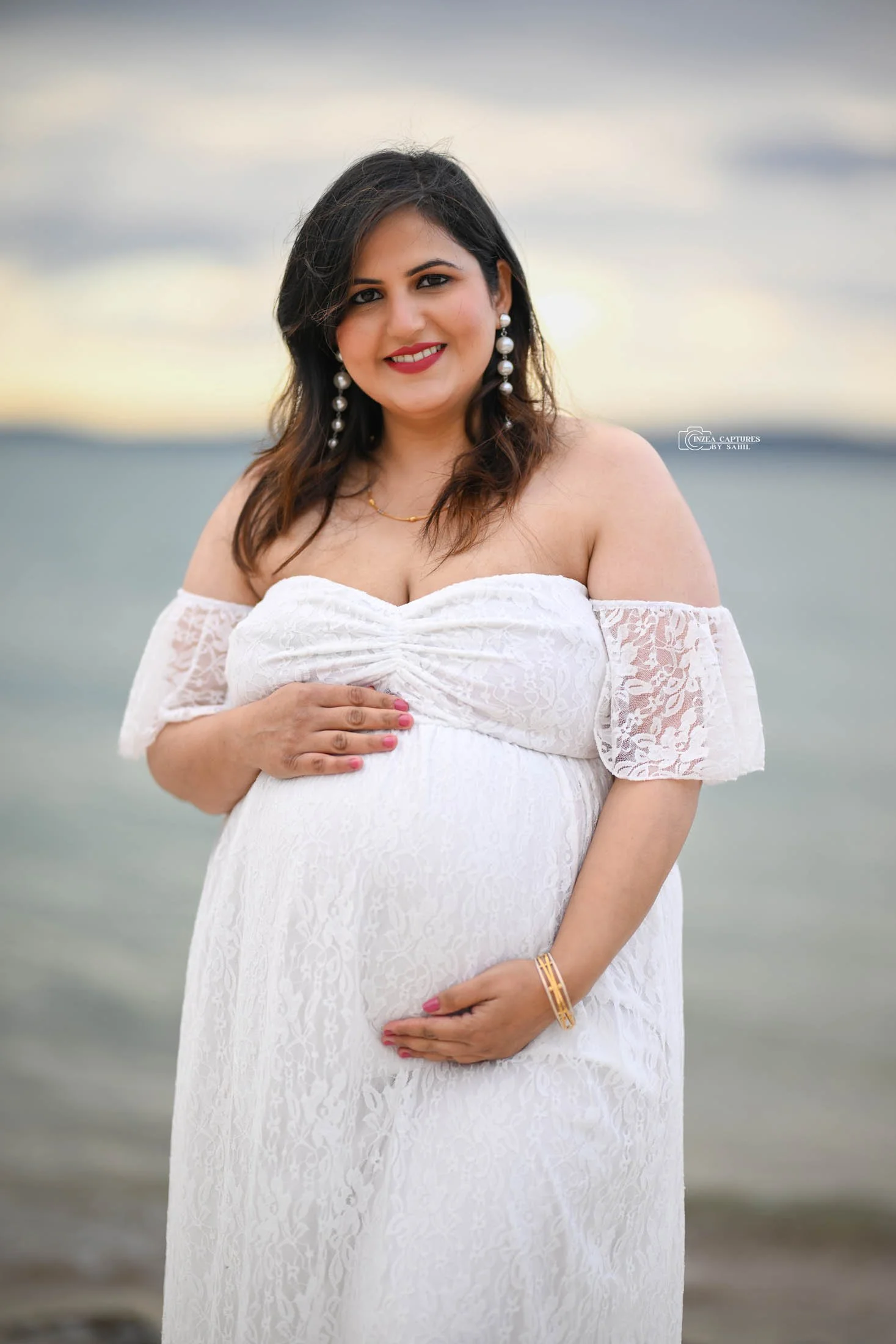 A pregnant woman is standing on a beach at sunset, wearing a white off-the-shoulder dress, large pearl earrings, a gold bracelet, and a gold necklace, smiling and holding her belly with one hand.