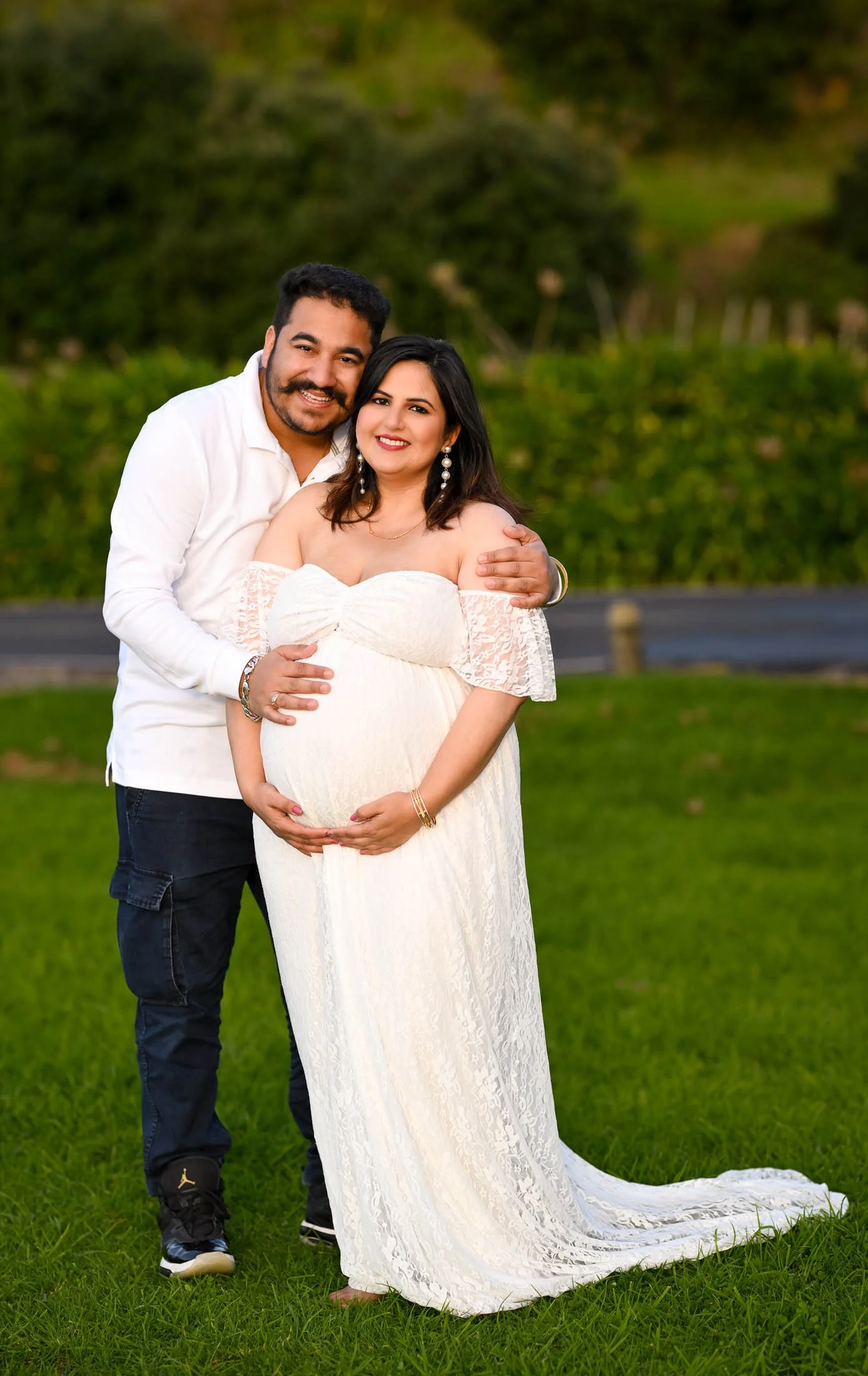 A smiling pregnant woman in a white lace dress and earrings standing on grass, with a man in a white shirt and dark jeans, smiling and wrapping his arm around her, in an outdoor park setting with trees and greenery in the background.