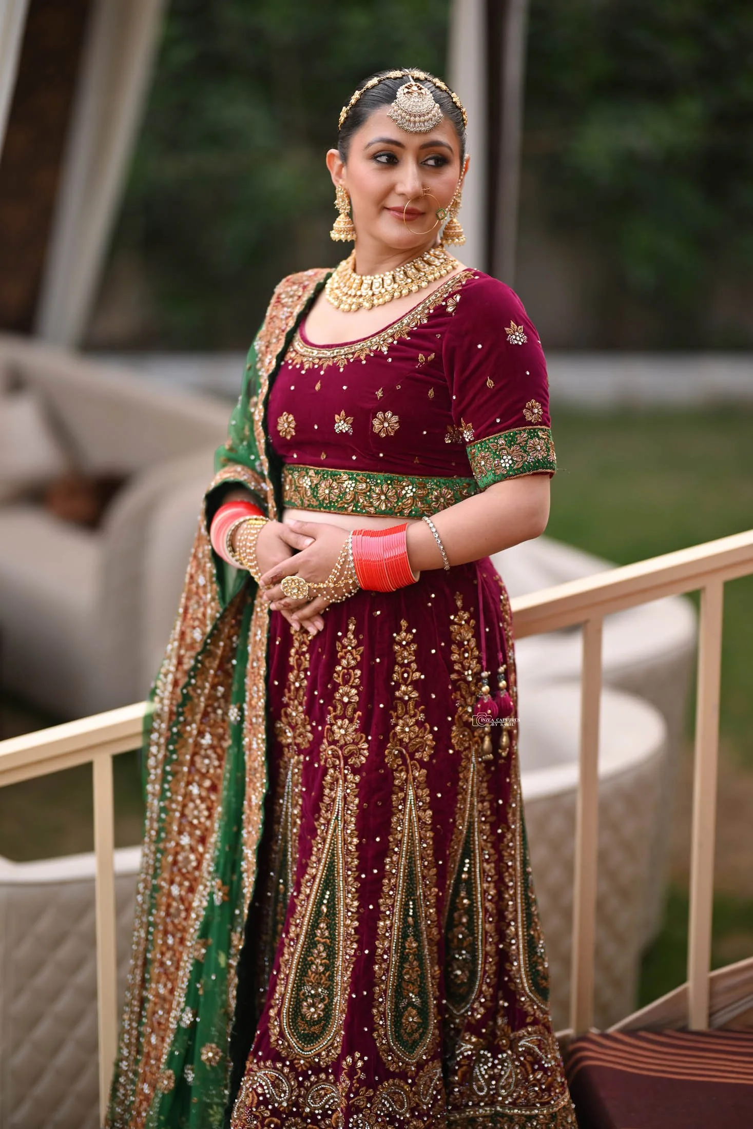 A woman wearing a traditional Indian outfit, a maroon and green embroidered lehenga with gold embellishments, gold jewelry, and red bangles, standing outdoors with greenery in the background.