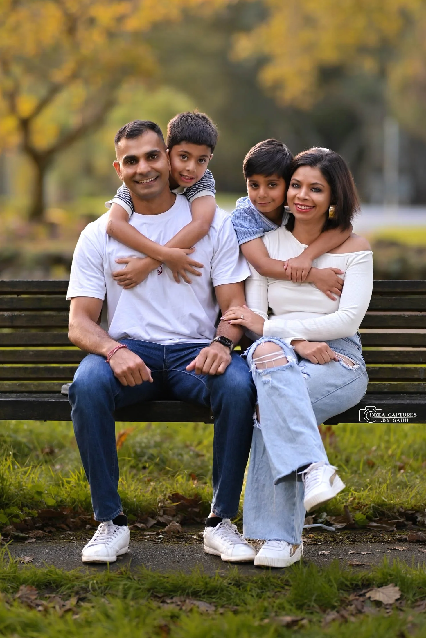 A family of four sitting on a park bench during autumn, smiling and embracing each other with trees and foliage in the background.
