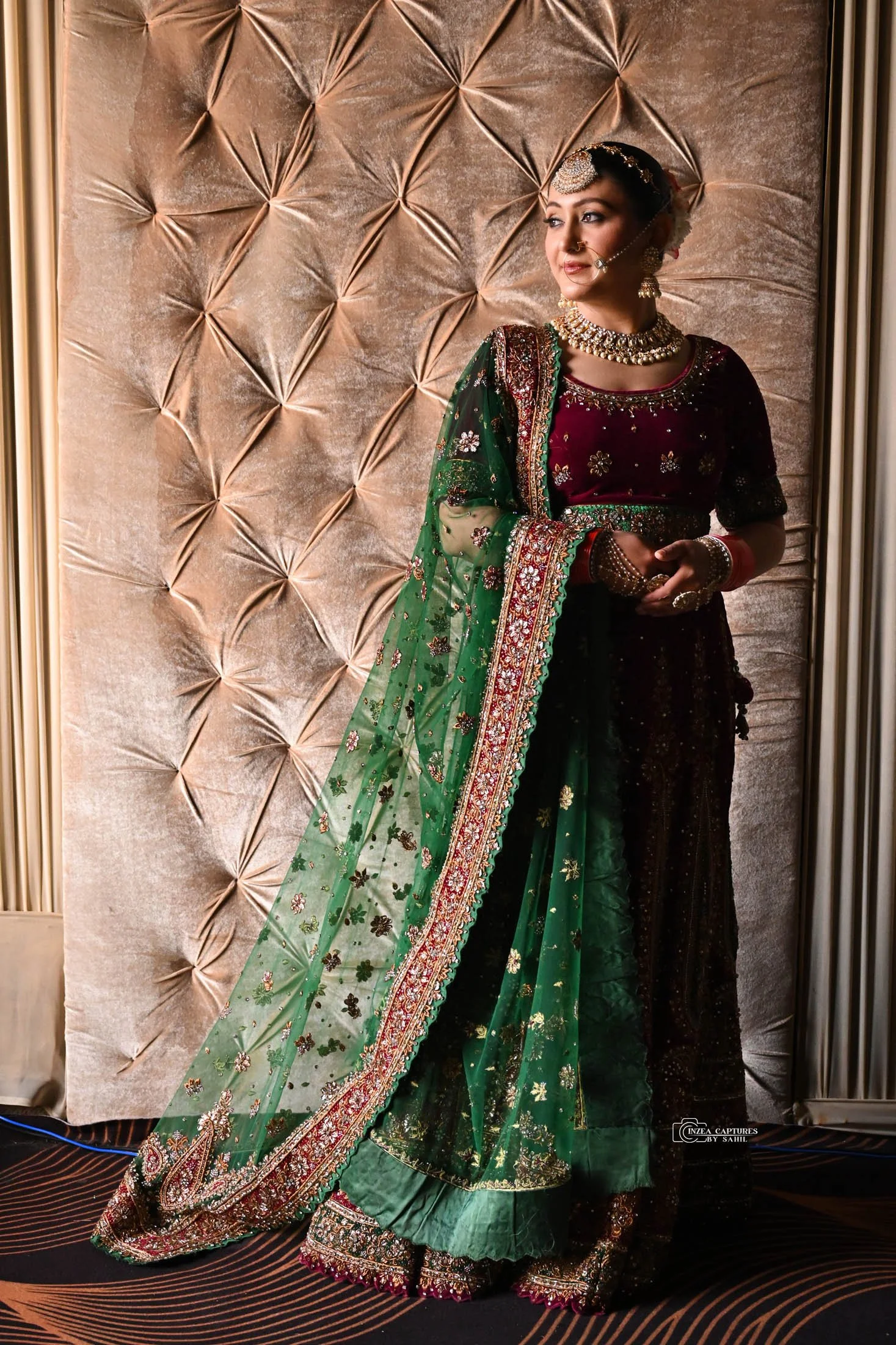 A woman dressed in traditional Indian bridal attire, wearing a green and maroon lehenga with intricate embroidery and jewelry, standing against a textured beige backdrop.
