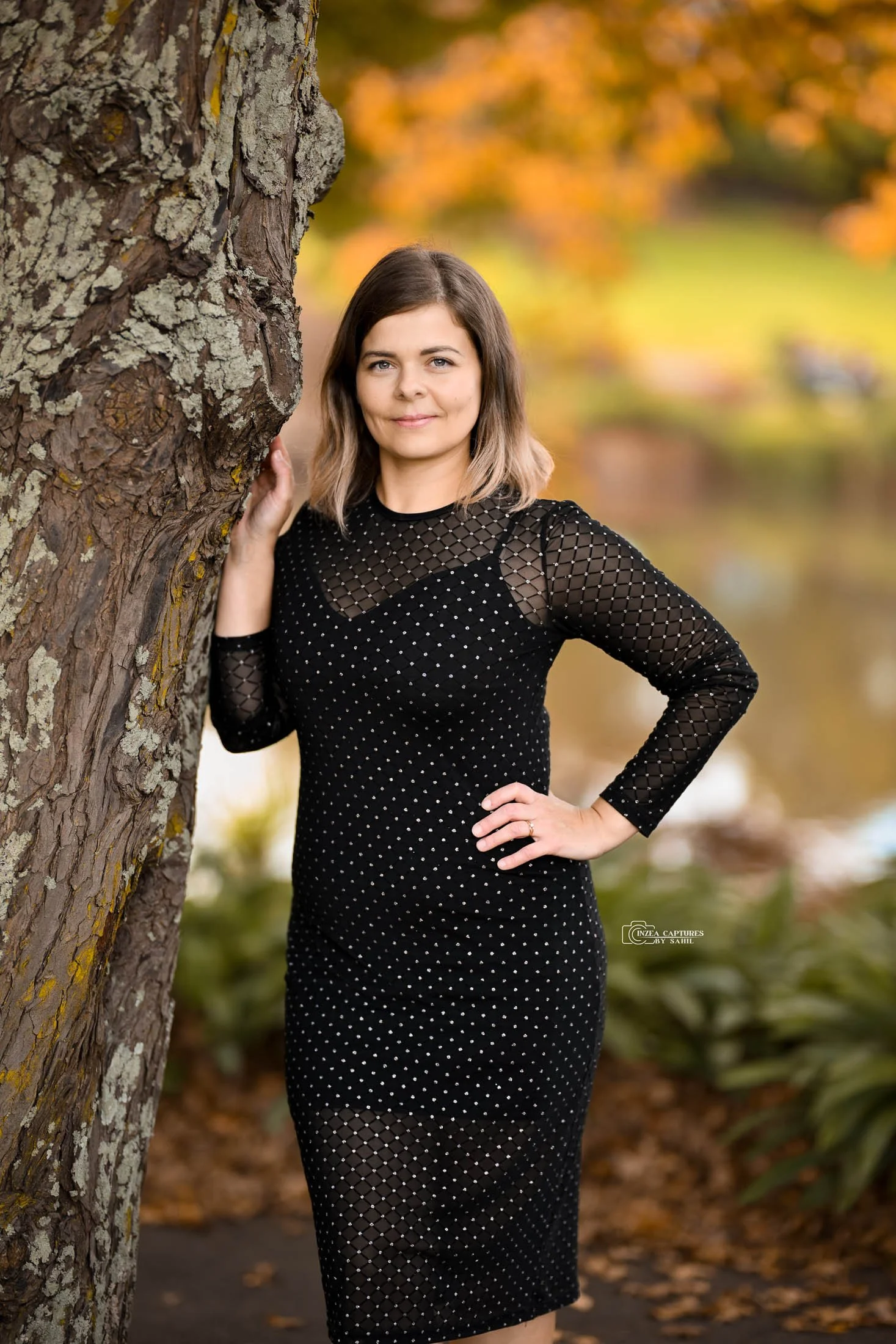 A woman in a black, mesh, polka-dot dress standing outdoors near a large tree with autumn-colored leaves in the background.