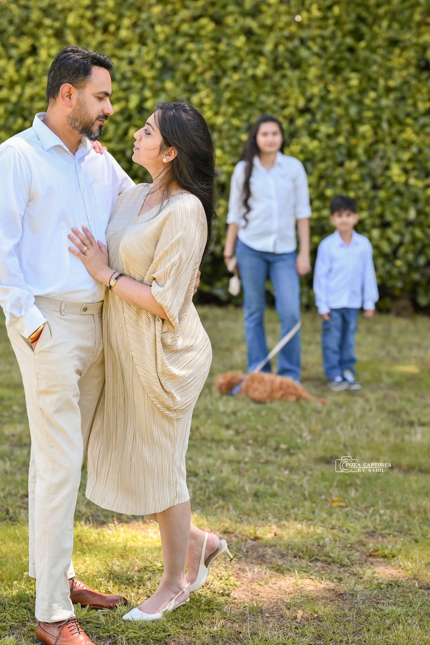 A couple facing each other and standing close, with two children and a dog in the background.