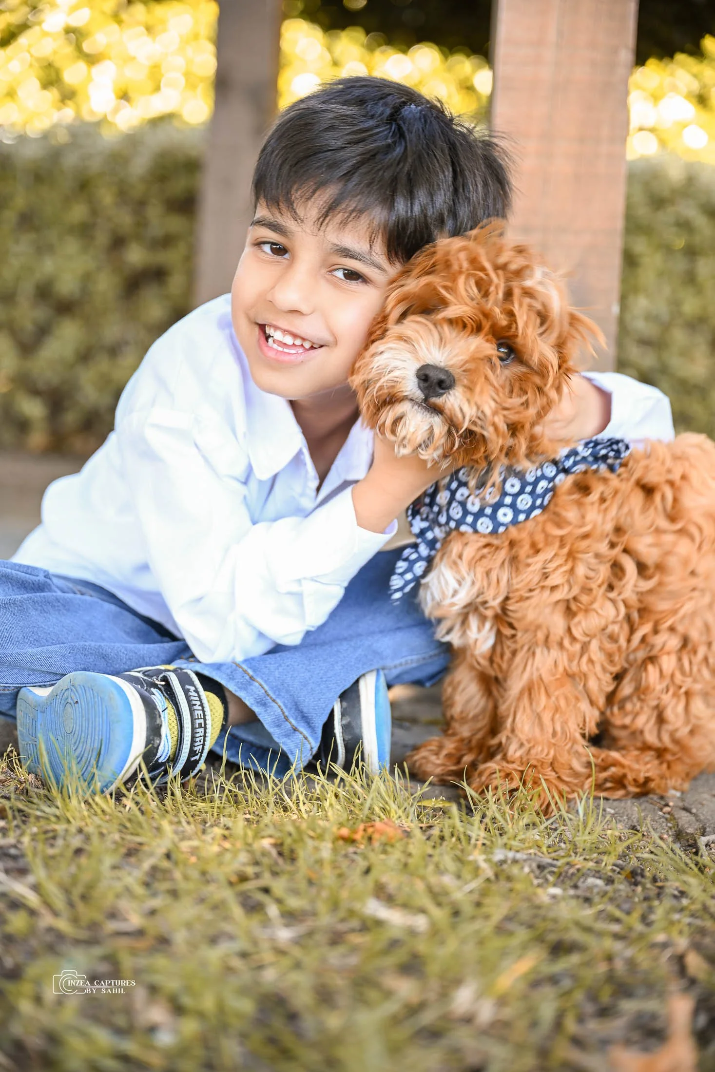 A young boy with dark hair smiling and hugging a curly-haired brown puppy outdoors on grass, with trees and sunlight in the background.