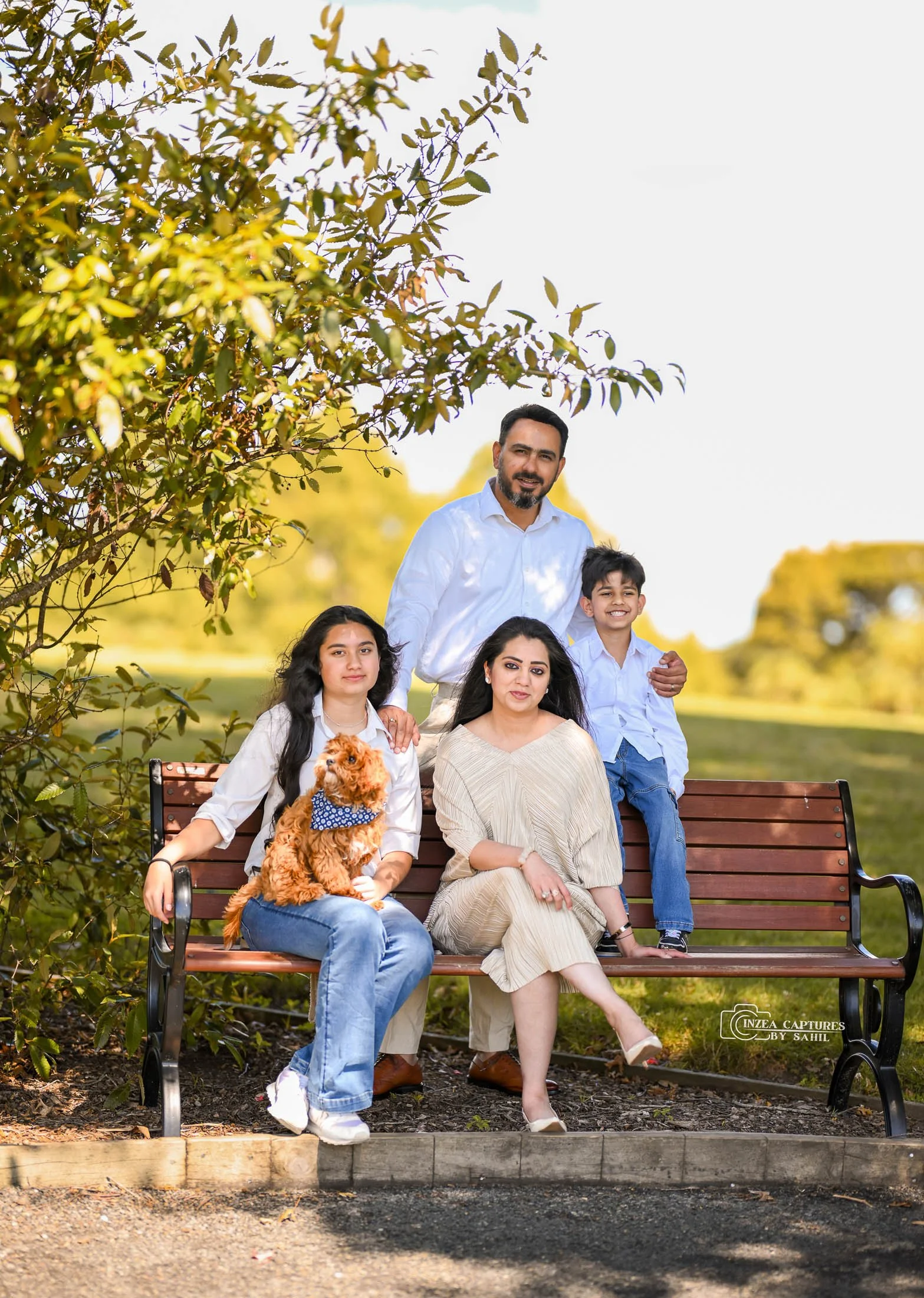 A family of four with a dog sitting on a park bench outdoors on a sunny day, smiling at the camera. The man and boy are standing behind the bench, while the woman and girl are seated. The girl holds a small, brown, curly-haired dog with a blue bandan
