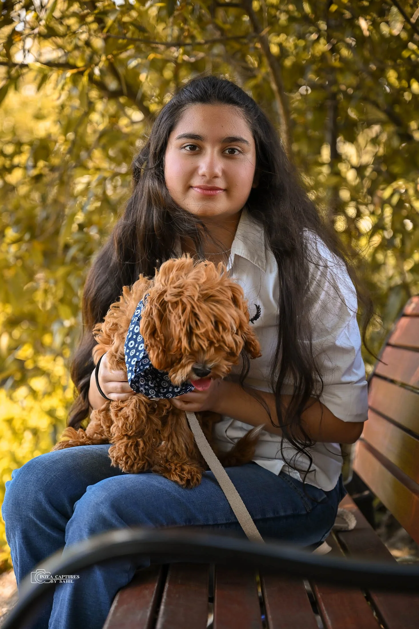 A girl with long dark hair sitting on a park bench holding a small brown dog wearing a blue bandana, surrounded by trees with autumn leaves.