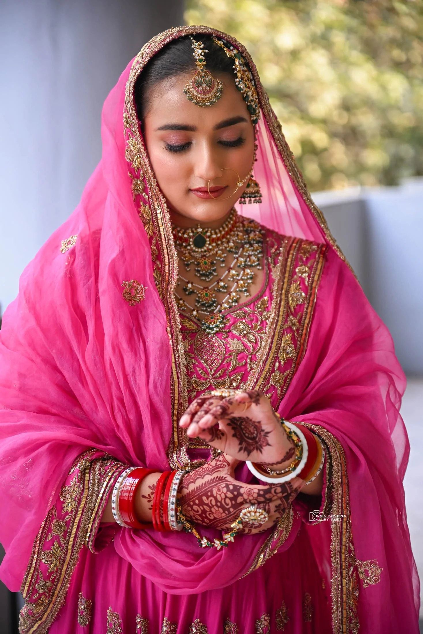 A bride dressed in a vibrant pink traditional Indian outfit with intricate gold embroidery, wearing elaborate jewelry including necklaces, earrings, and a headpiece, with henna designs on her hands, looking downward.