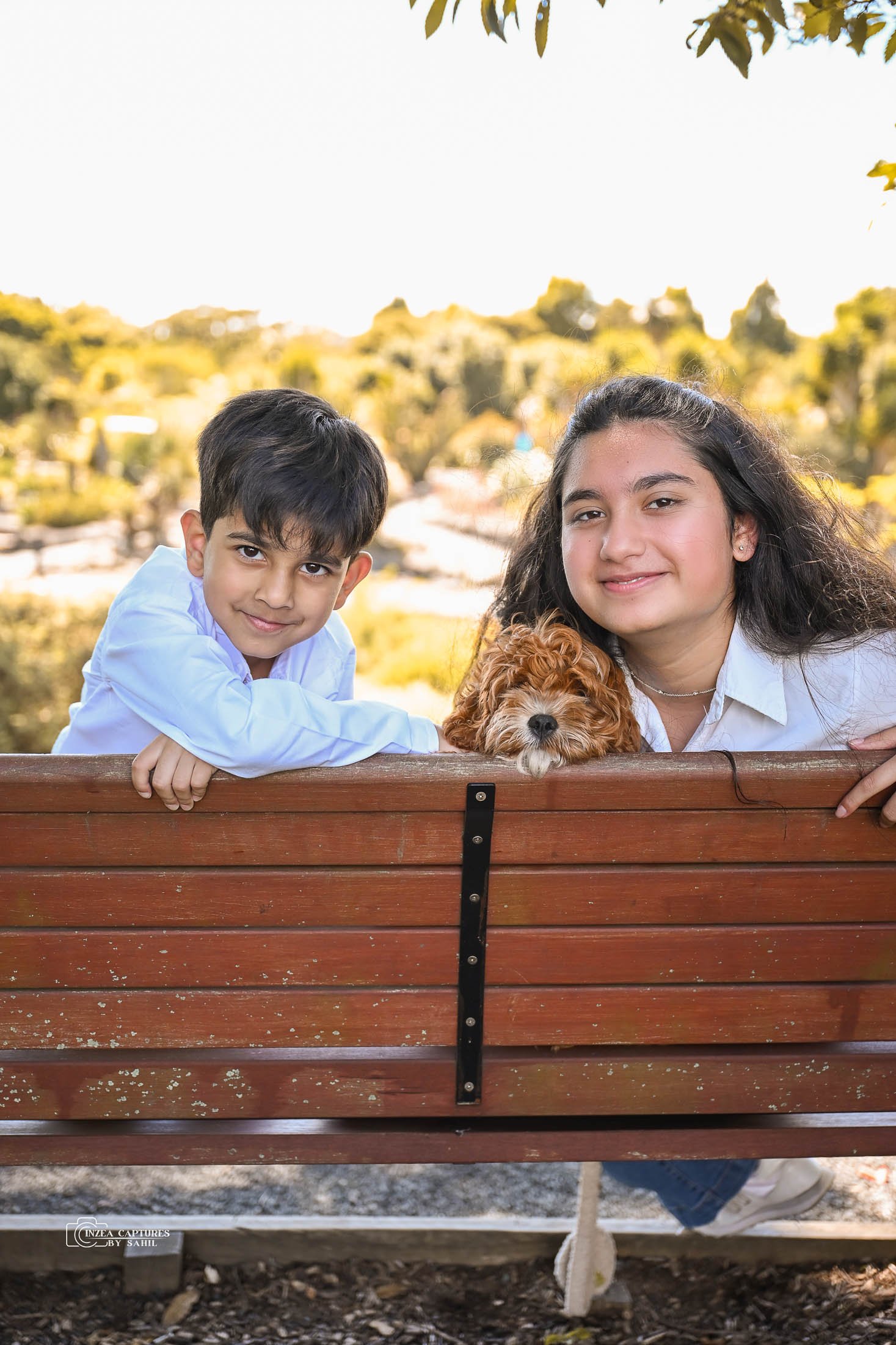 Two children, a boy and a girl, leaning over a wooden park bench with a small brown dog between them, outdoors in a park with trees and clear sky in the background.