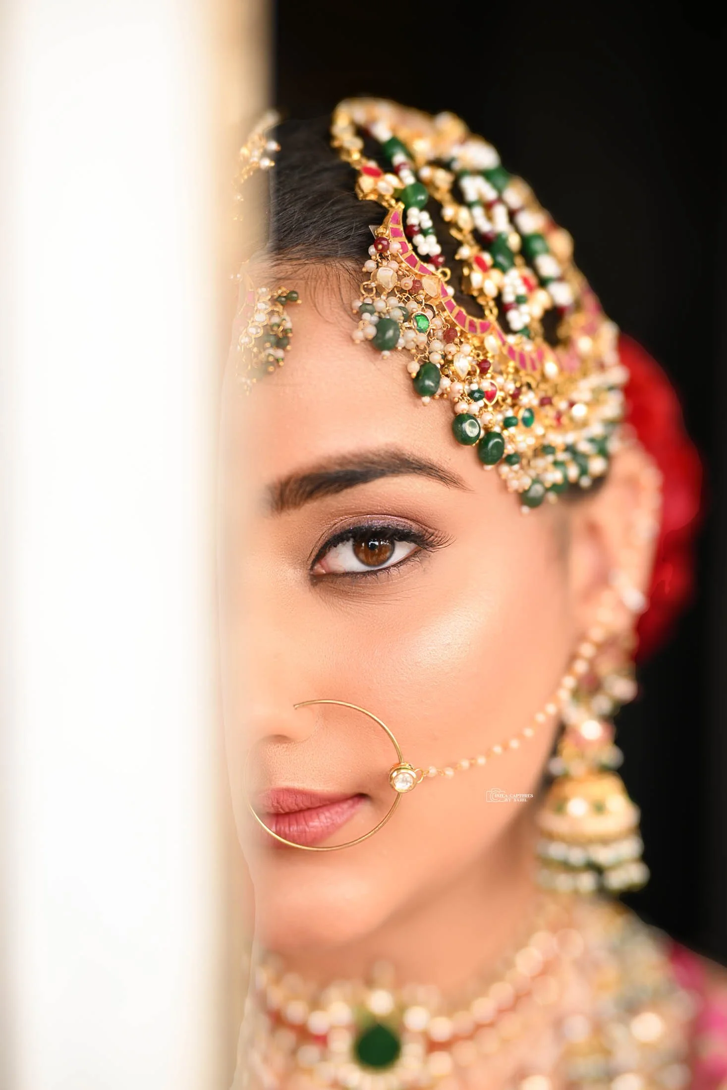 A close-up of a woman wearing traditional Indian jewelry, including a large nose ring connected to her ear, a decorated headpiece with pearls, green gemstones, and gold accents, and matching earrings and necklace.