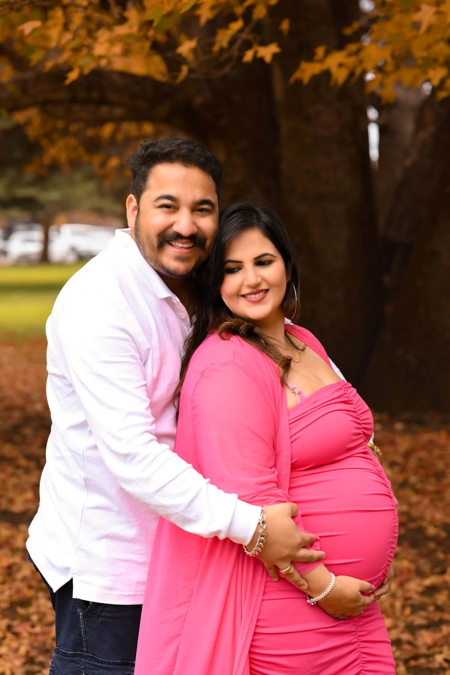 A couple outdoors in autumn, with the man standing behind the pregnant woman, both smiling. The woman is wearing a pink dress and the man is in a white shirt. The background features a large tree with fall foliage.