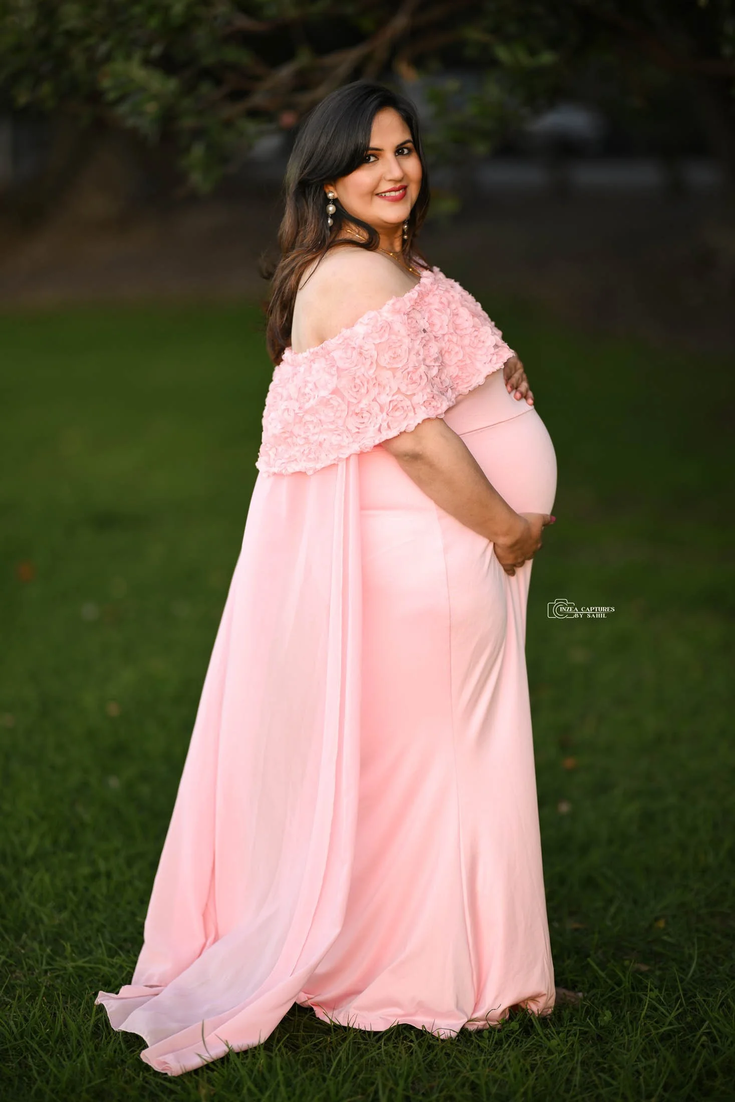 A pregnant woman in a pink gown and floral cape standing outdoors on grass, smiling and holding her belly.