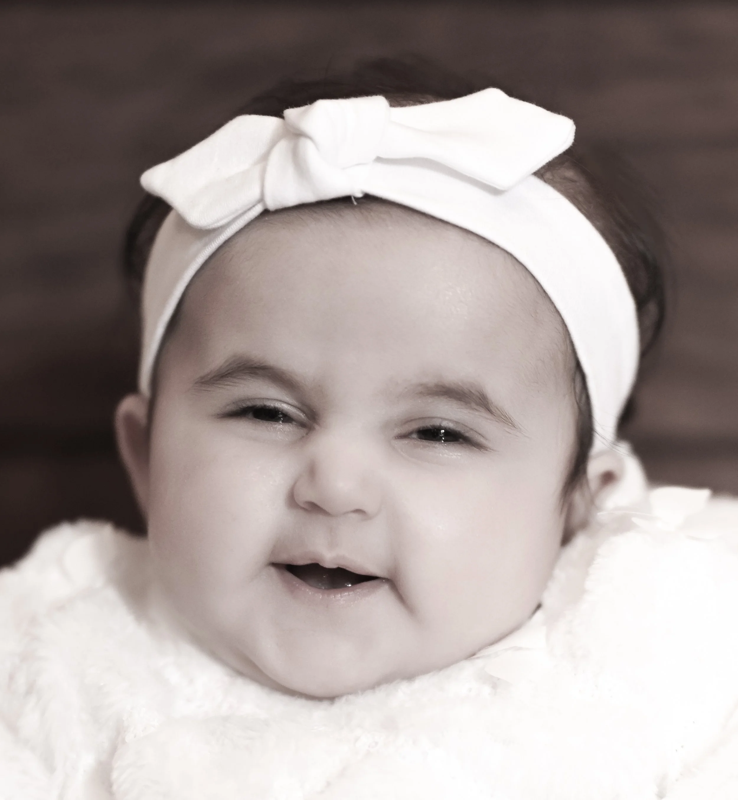 Close-up of a smiling baby girl wearing a white headband with a bow and a white fluffy outfit.