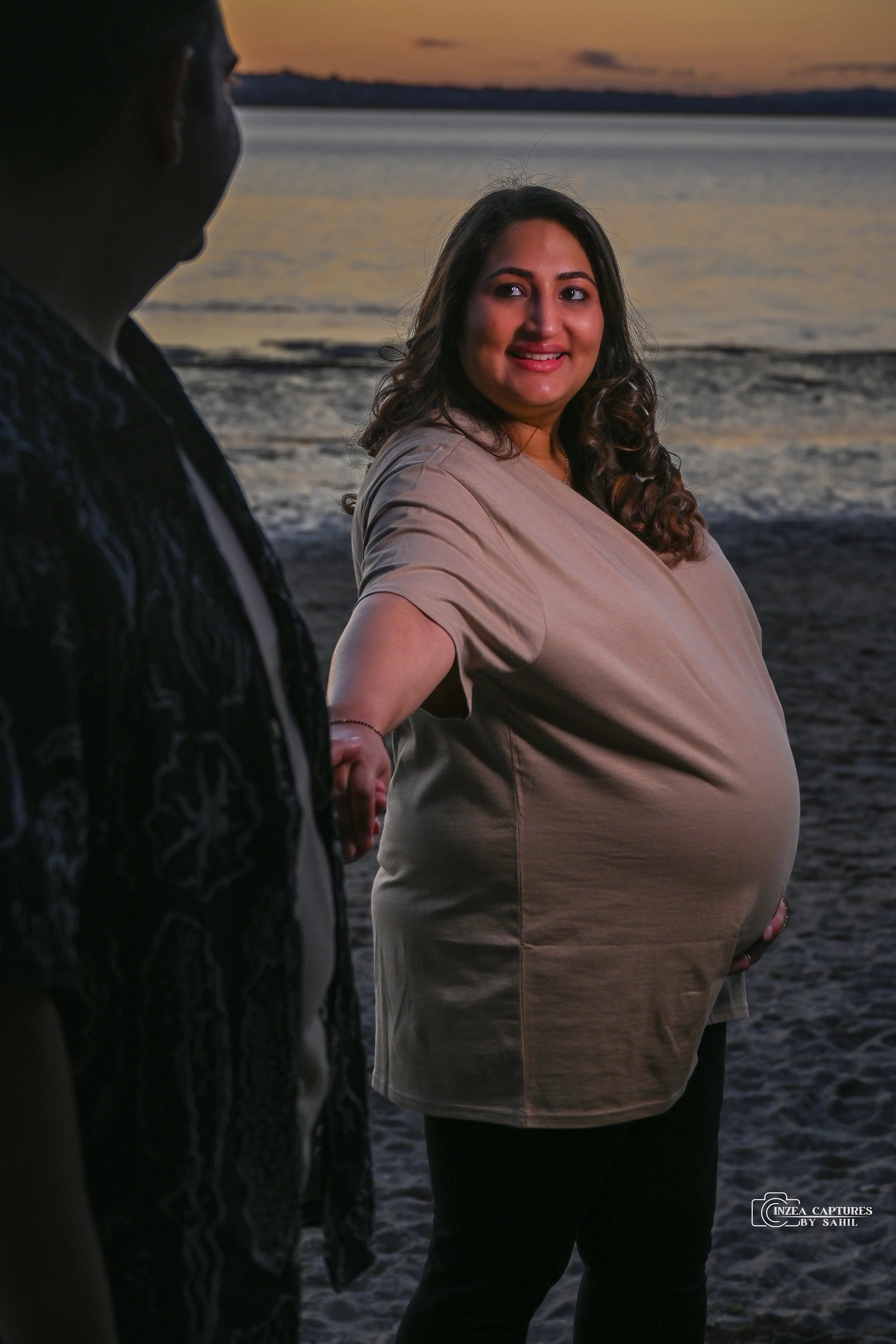 A pregnant woman smiling at a man holding her hand on the beach during sunset.