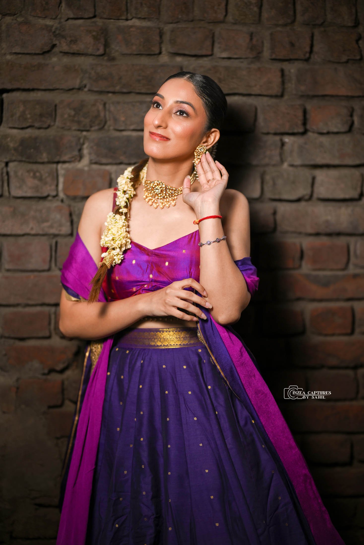 A woman wearing traditional Indian attire and jewelry, standing against a brick wall, touching her earring and looking upward.