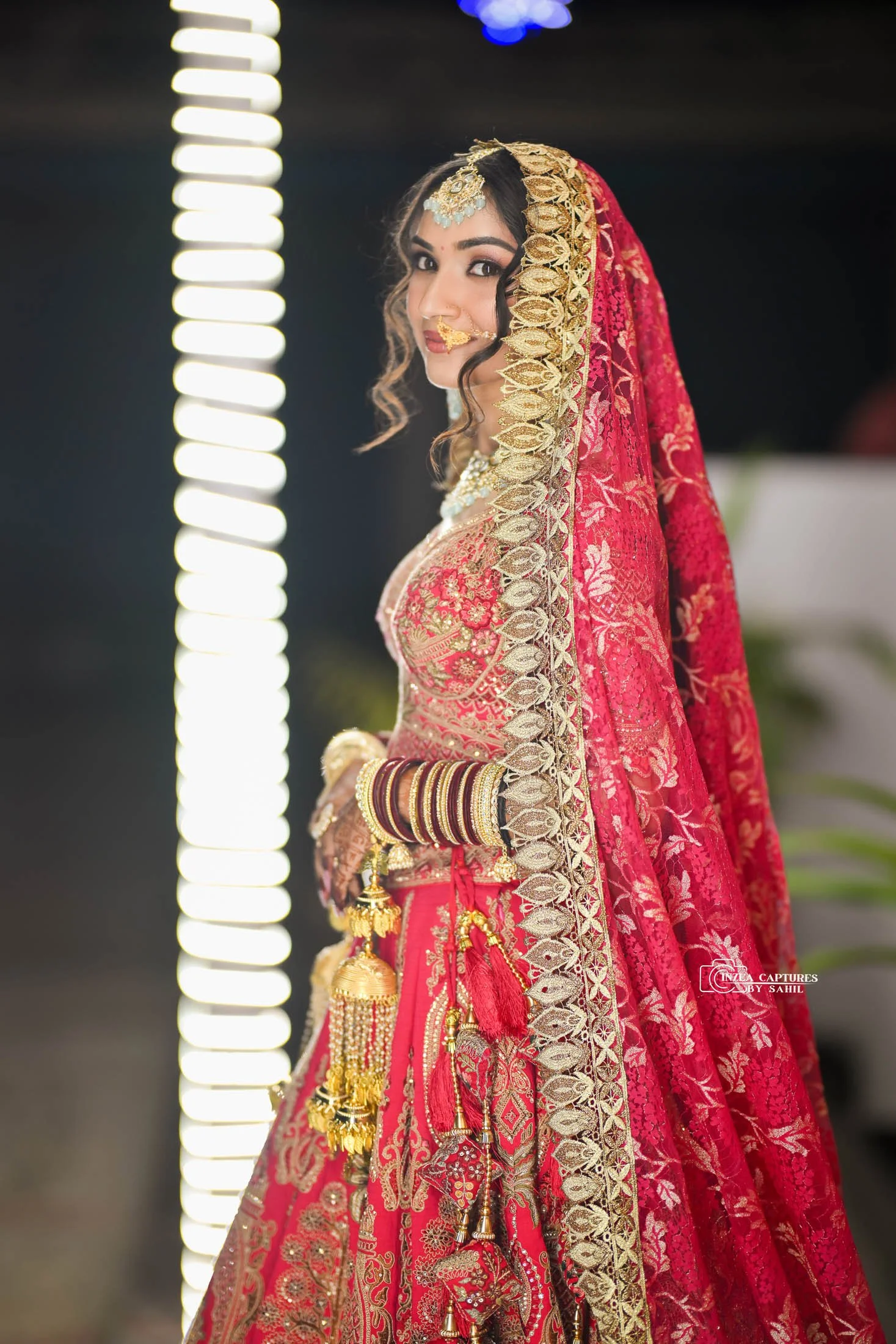 A woman dressed in traditional Indian bridal attire, wearing a red and gold embroidered saree with matching jewelry, bangles, and a head covering with intricate gold details, looking over her shoulder.