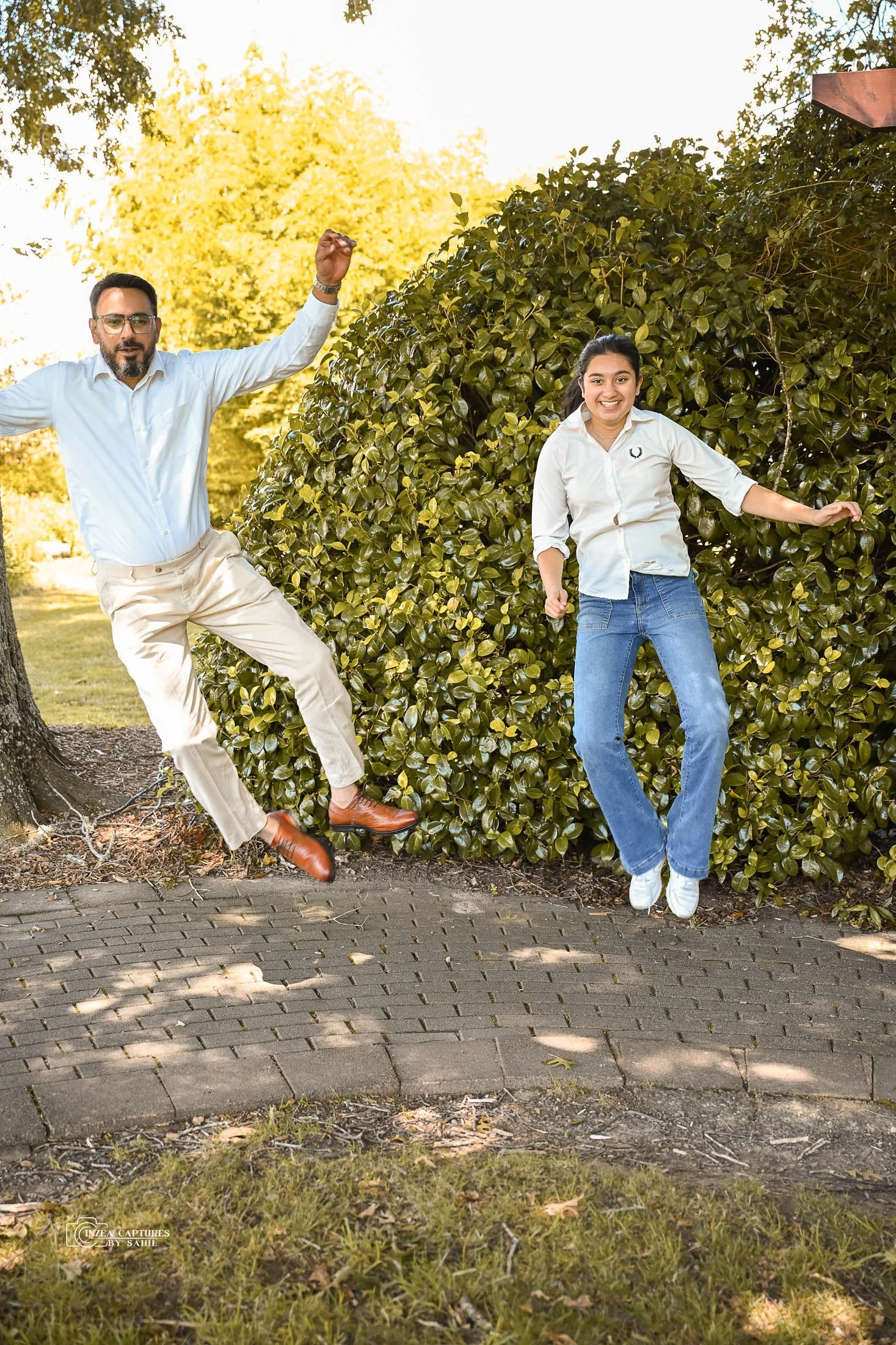 A man and a woman are jumping in the air outdoors on a sidewalk in front of a bush, with trees in the background. The man is wearing a white shirt, beige pants, and brown shoes, and the woman is wearing a white shirt, blue jeans, and white shoes. The
