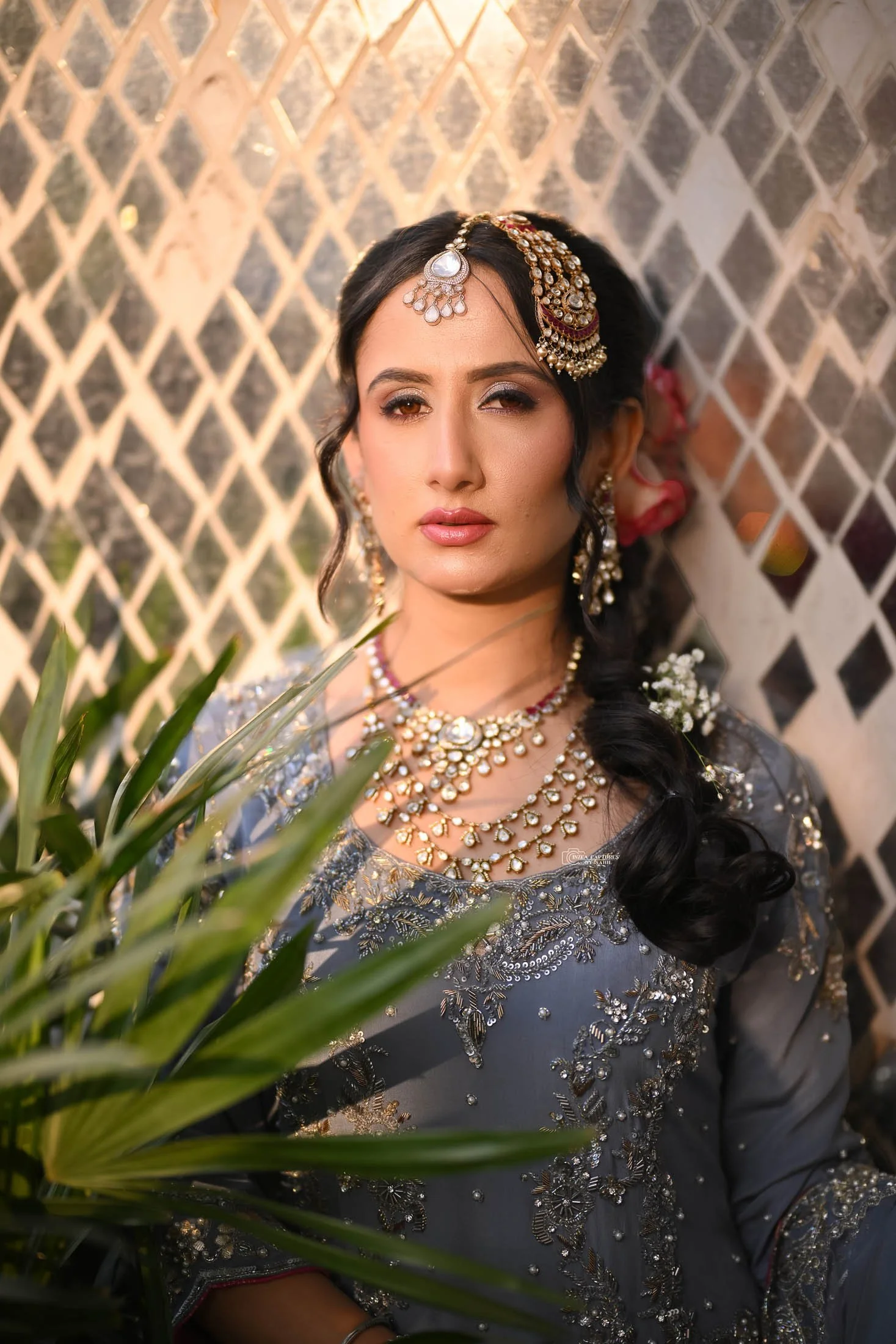 A woman dressed in traditional South Asian bridal attire, wearing elaborate jewelry, including earrings, necklaces, and a headpiece, with dark wavy hair and natural makeup, standing in front of a lattice fence with sunlight highlighting her face.