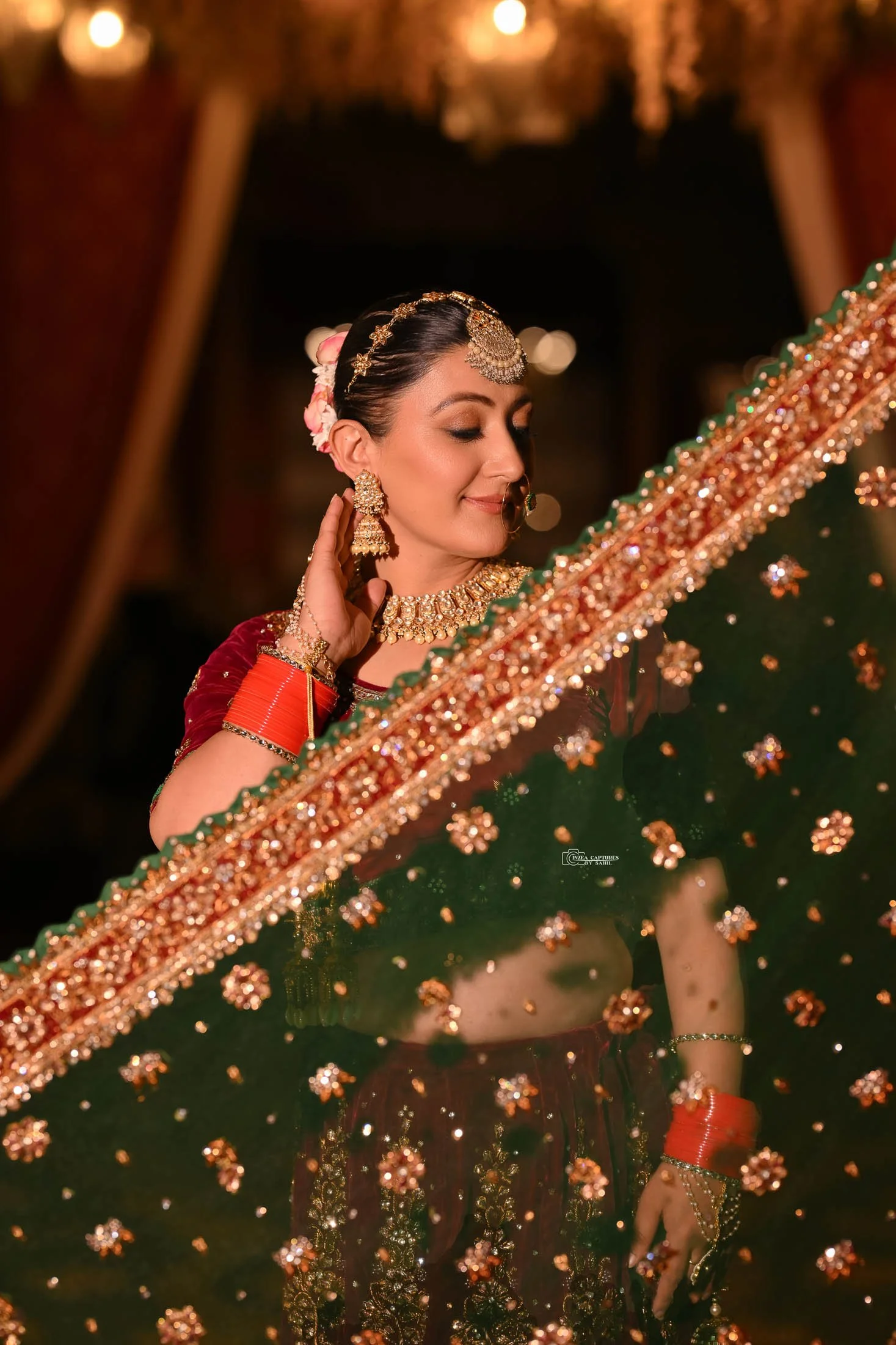 A woman dressed in traditional Indian attire and jewelry, standing indoors with warm lighting. She is looking down with a gentle expression, surrounded by ornate decorations.