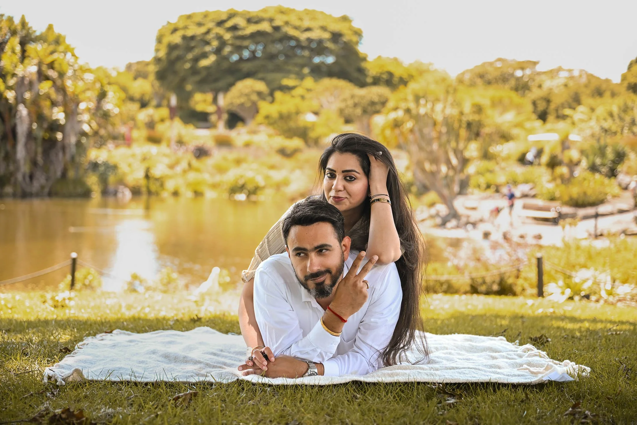 A couple relaxing on a blanket by a pond in a park with lush green trees in the background.