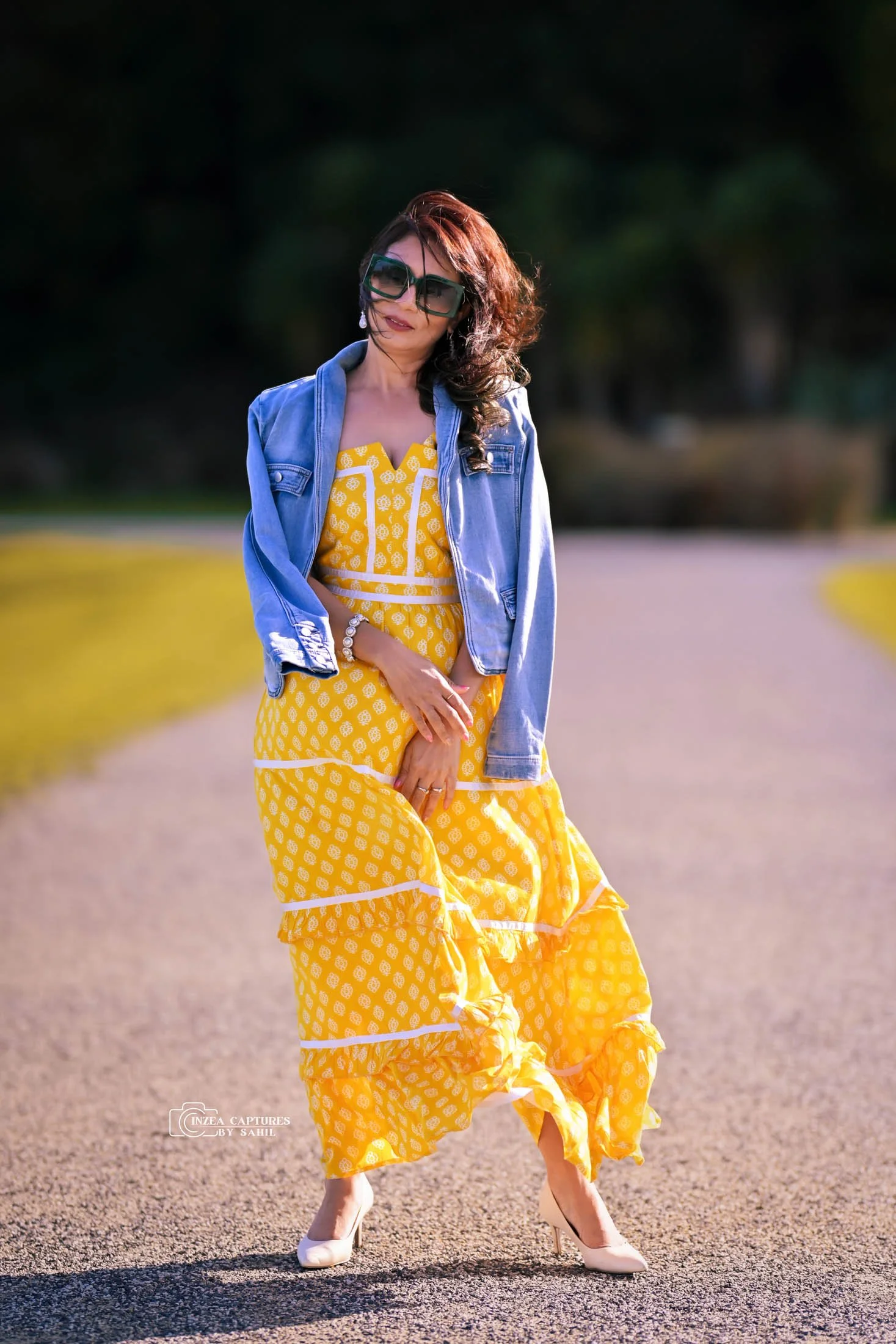 A woman wearing a yellow patterned maxi dress, a blue denim jacket, large sunglasses, and white high heels, standing outdoors on a gravel path with green trees in the background.