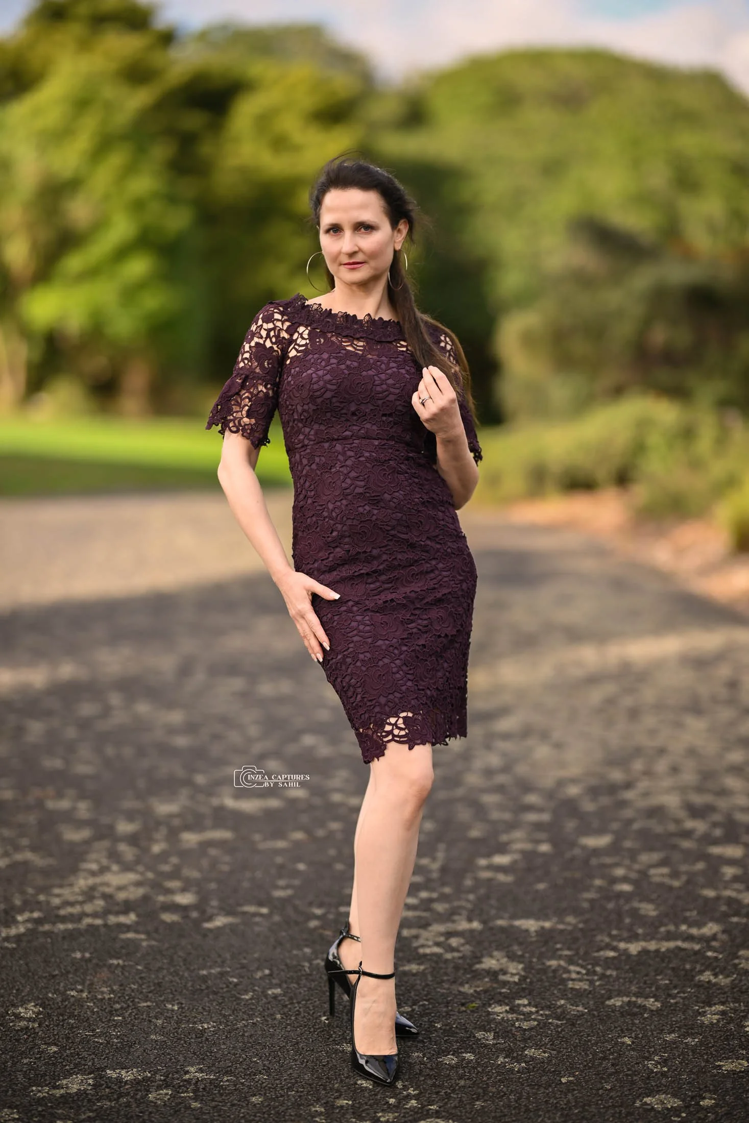 Woman in a dark purple lace dress with black heels standing outdoors on a paved path surrounded by green trees.