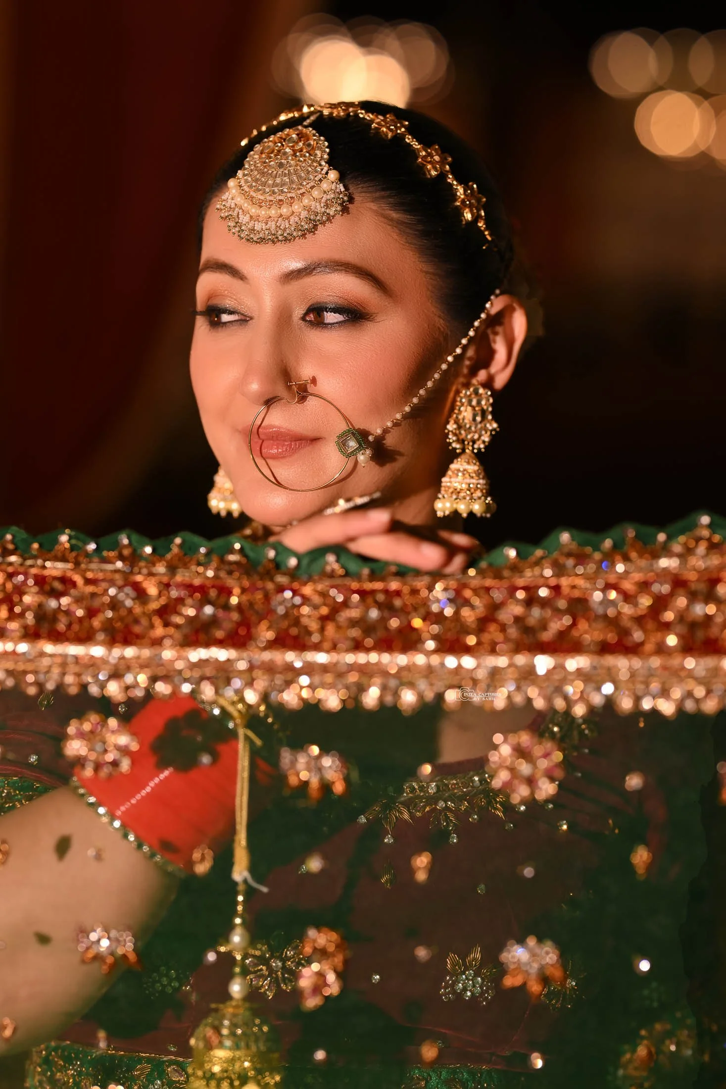 A woman dressed in traditional Indian jewelry, including earrings, a headpiece, nose ring, and bangles, with a soft, blurred background and warm lighting.