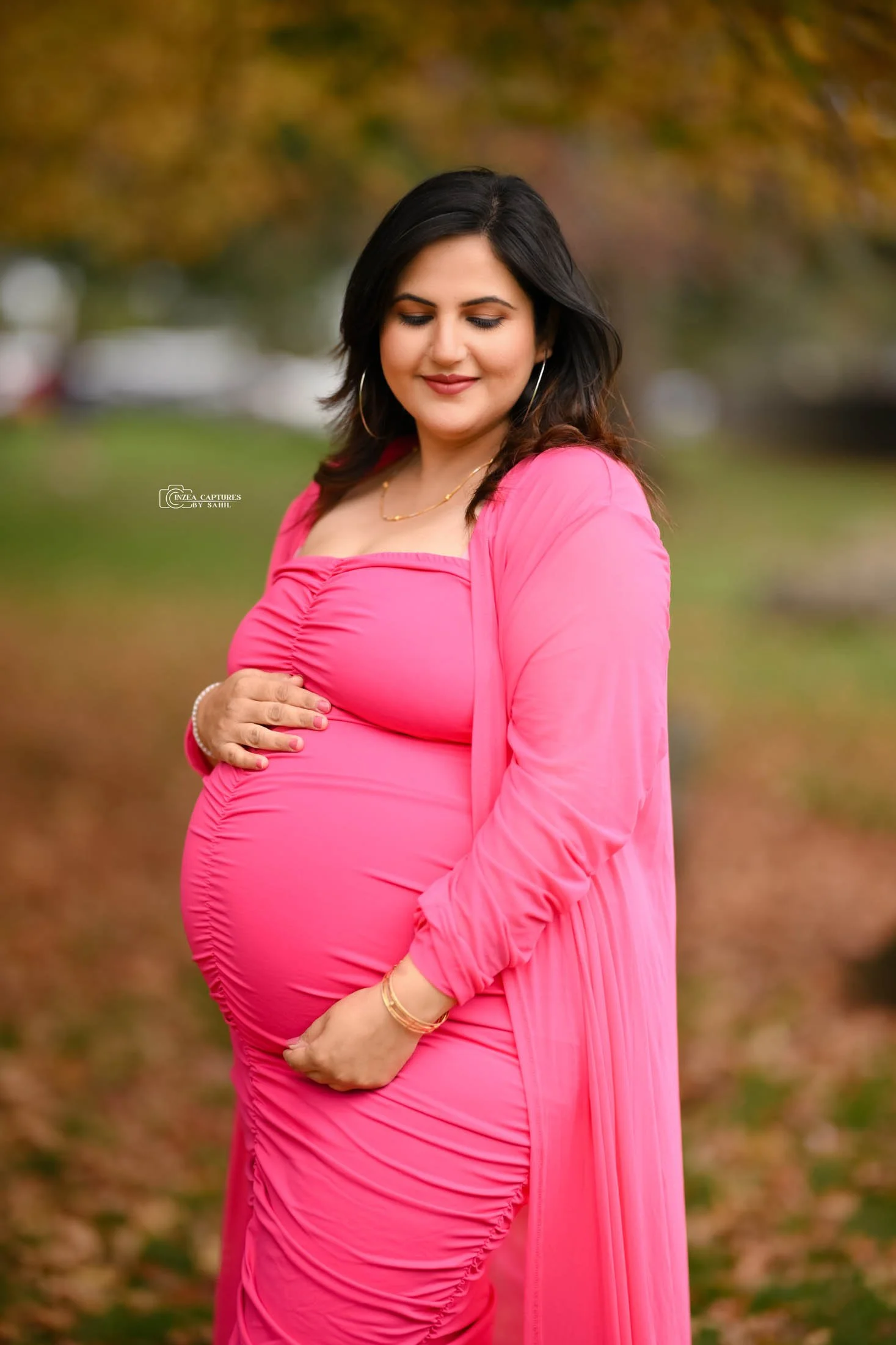 A pregnant woman in a pink dress and matching cover-up standing outdoors on fallen autumn leaves, with trees and blurred background.