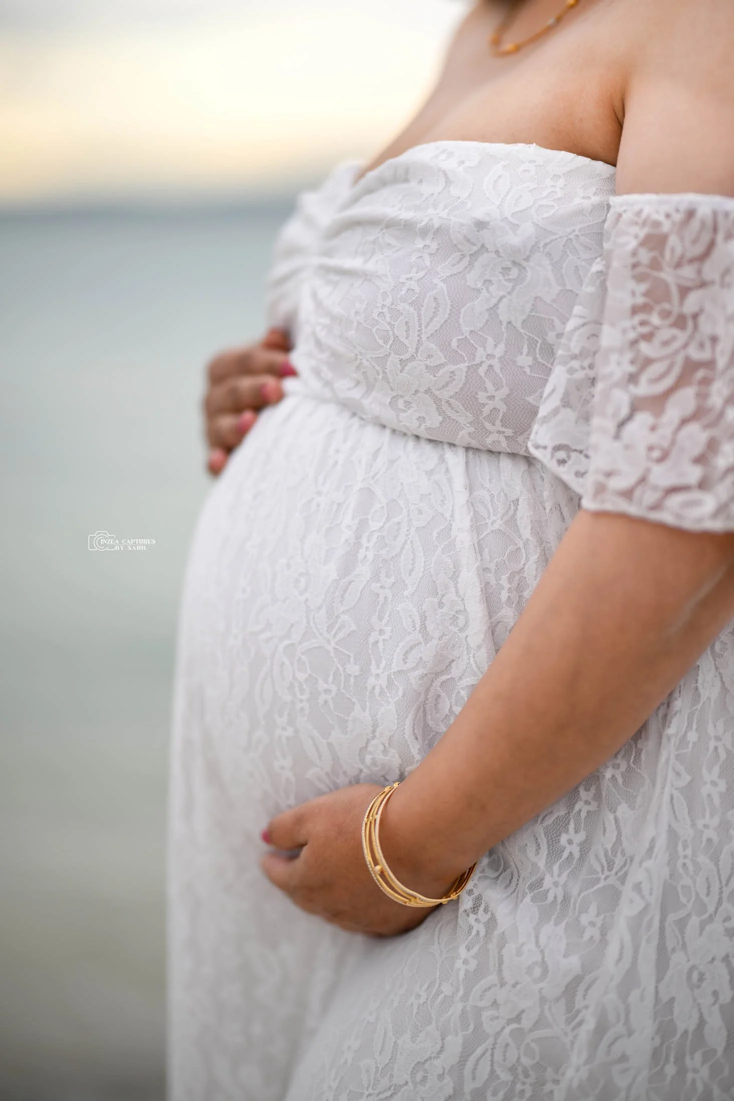 Close-up of a pregnant woman wearing a white lace dress, holding her belly with one hand, and wearing gold bangles and a gold necklace.