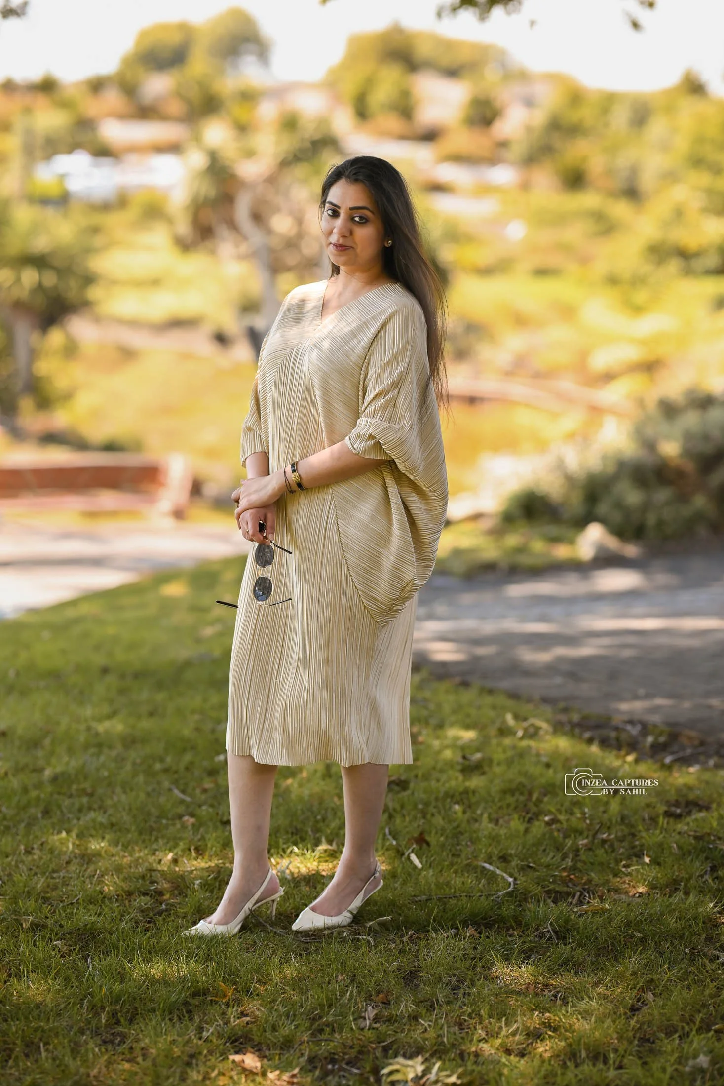 A woman in a gold, pleated dress standing outdoors on grass, holding sunglasses in her right hand, with a blurred background of trees and a park.