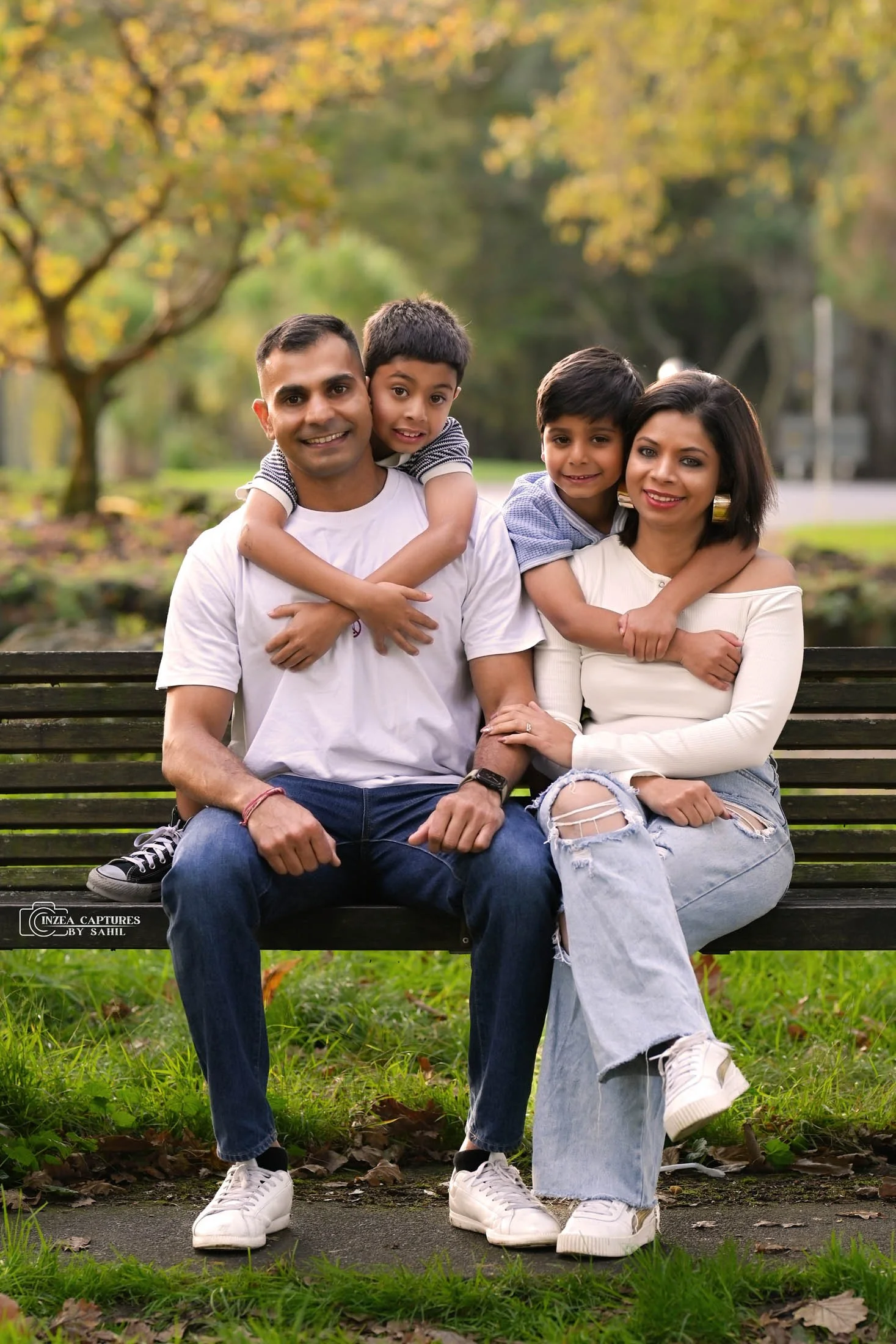 A smiling family of four sitting on a park bench; two children hugging their parents, in a park with trees showing fall foliage.