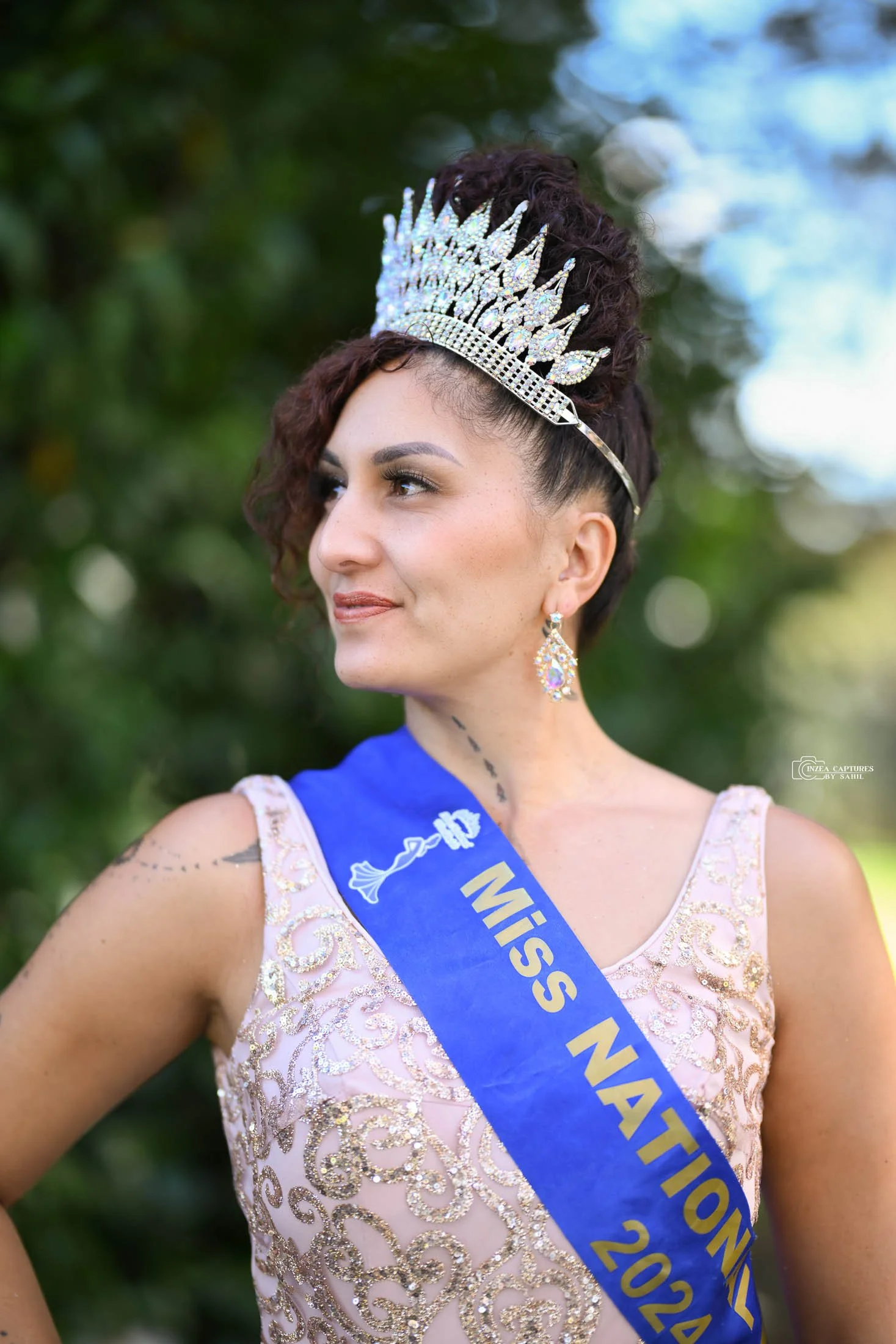Miss Nation 2024 woman wearing a crown and sash, dressed in a light pink dress with gold embellishments, with a background of green trees.