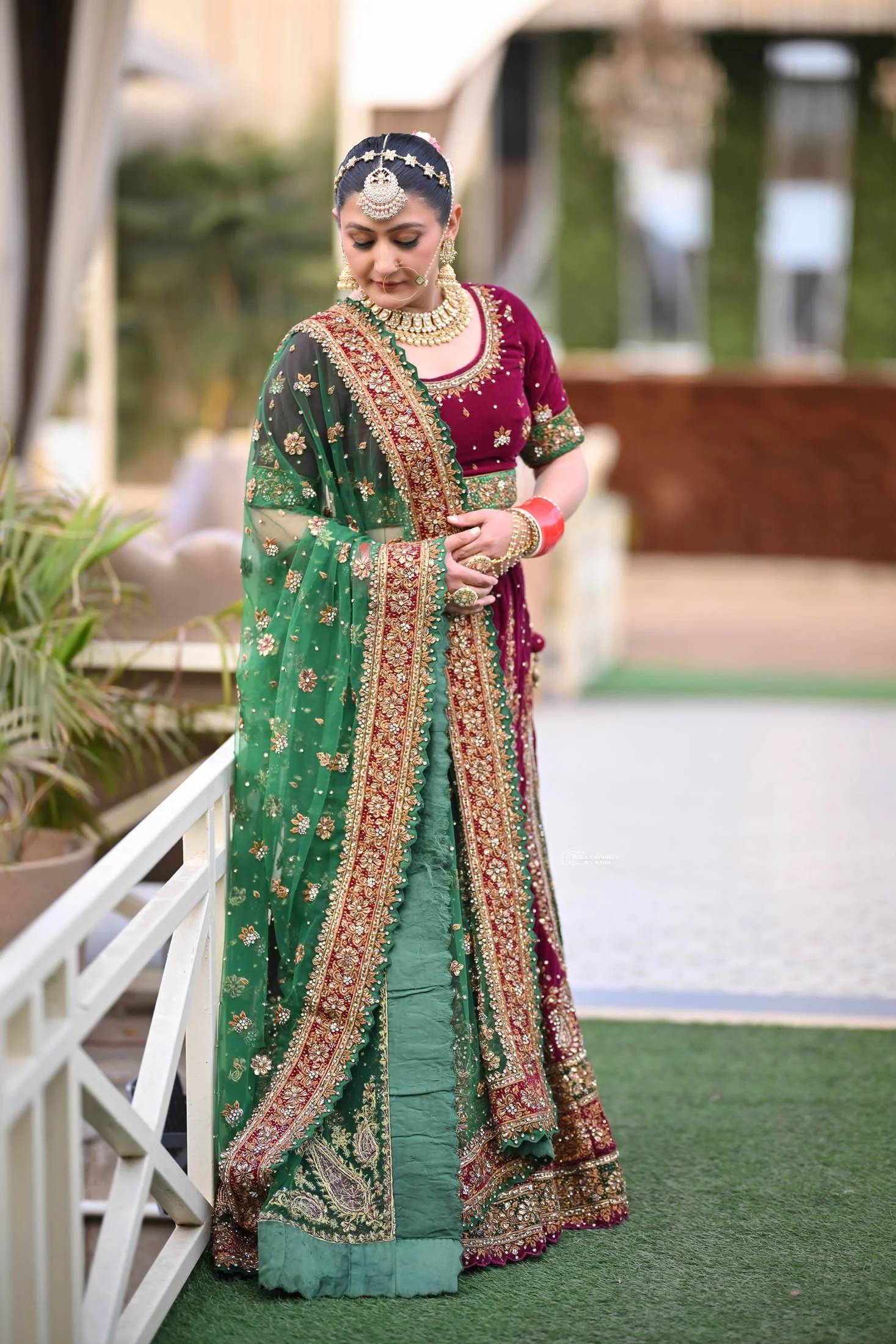 A woman dressed in traditional Indian bridal attire, wearing a richly embroidered maroon and green saree, with heavy jewelry including a necklace, earrings, headpiece, and bangles, standing outdoors on a green carpet.