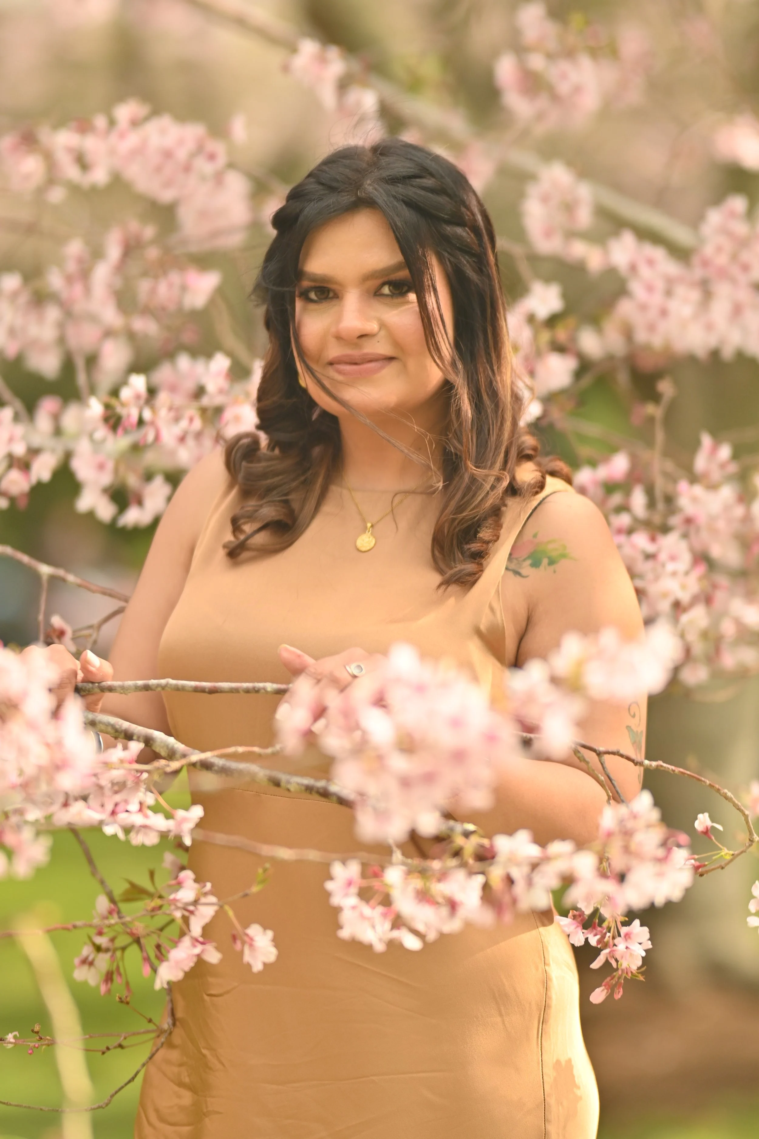 A woman standing among blooming pink cherry blossom branches, wearing a tan dress and a gold necklace, with a slight smile.