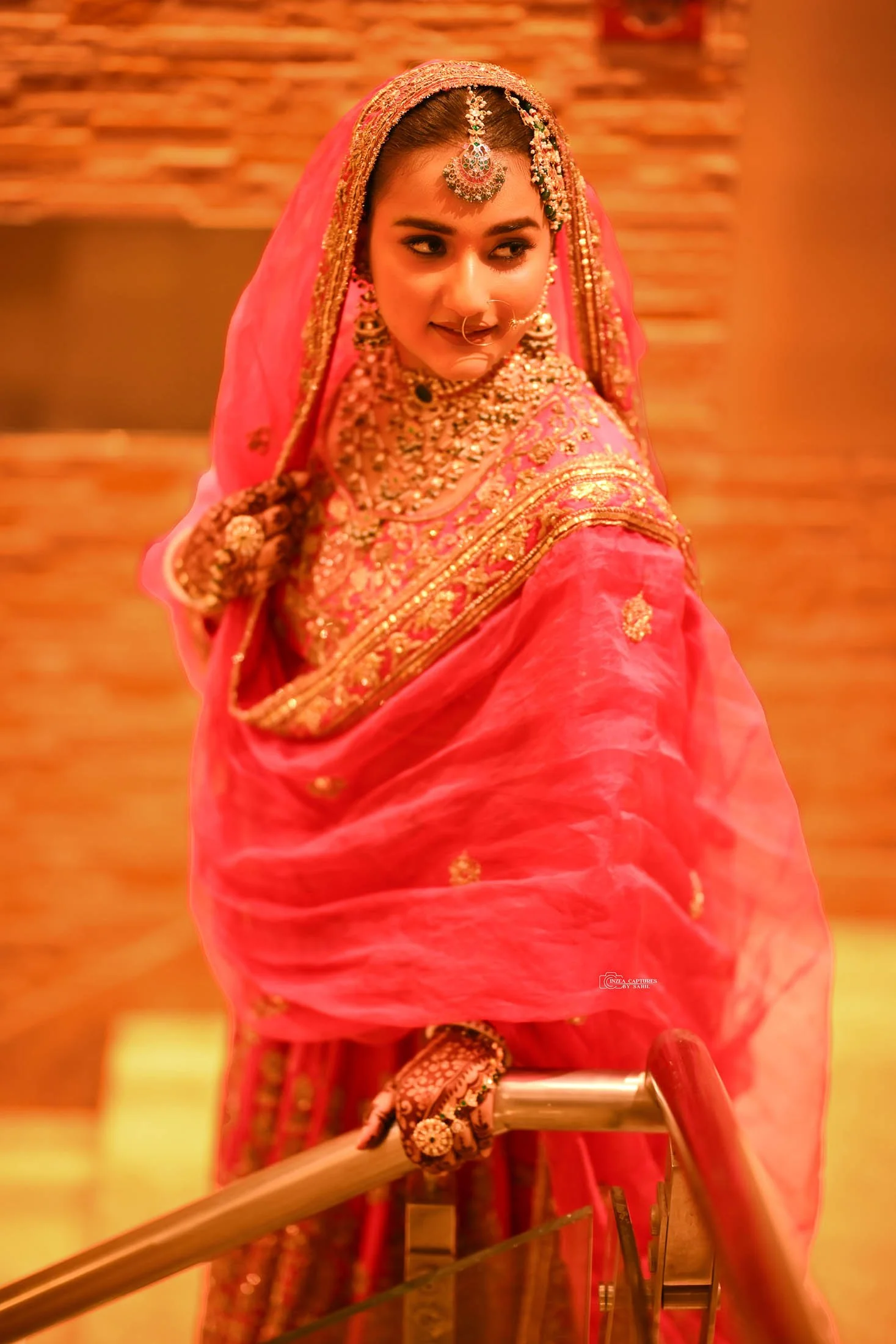 A woman in traditional Indian bridal attire, wearing a pink saree with gold embroidery, jewelry, and a nose ring, looking over her shoulder.