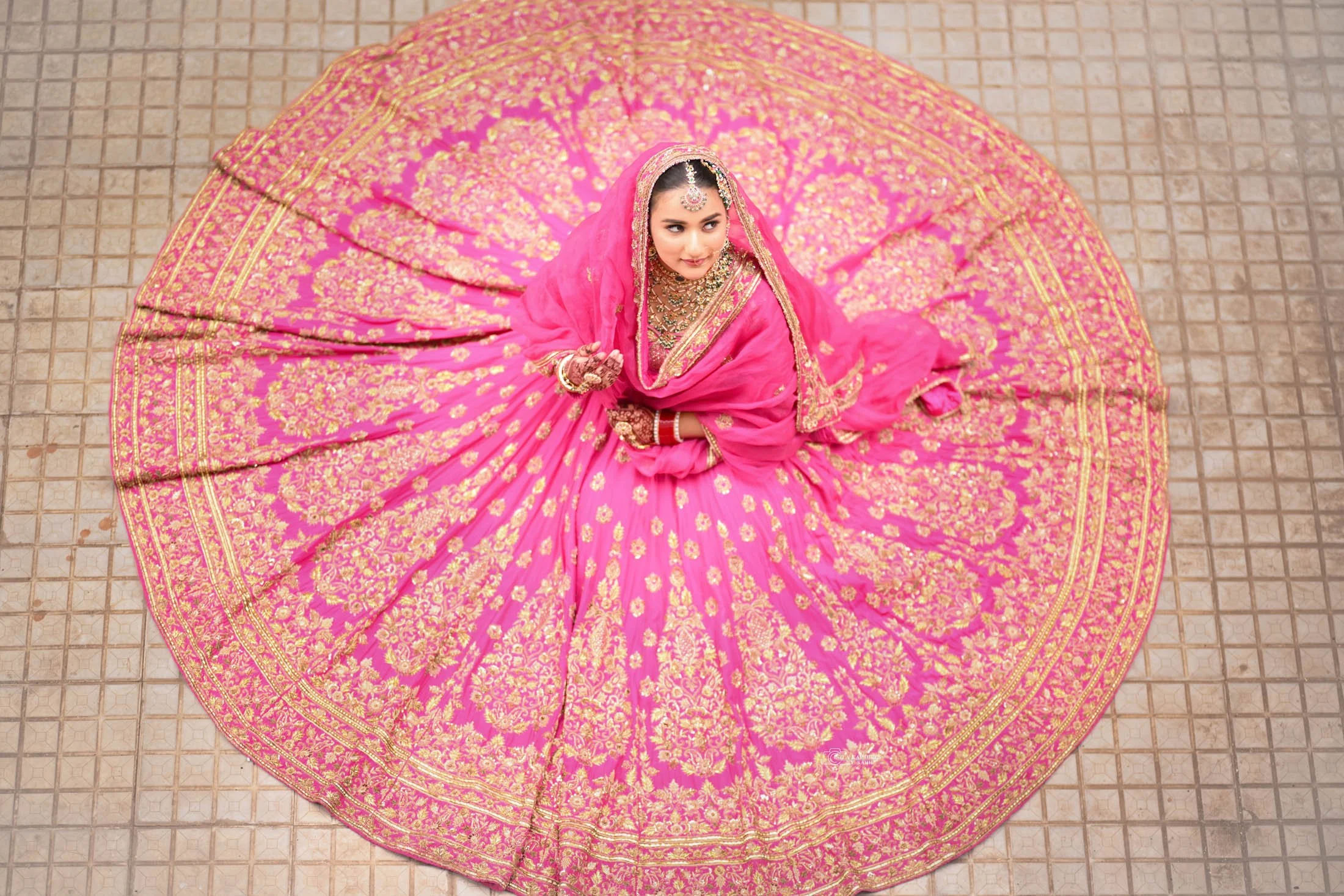 A woman in a pink and gold bridal lehenga with intricate embroidery, sitting on the tiled floor, looking up.