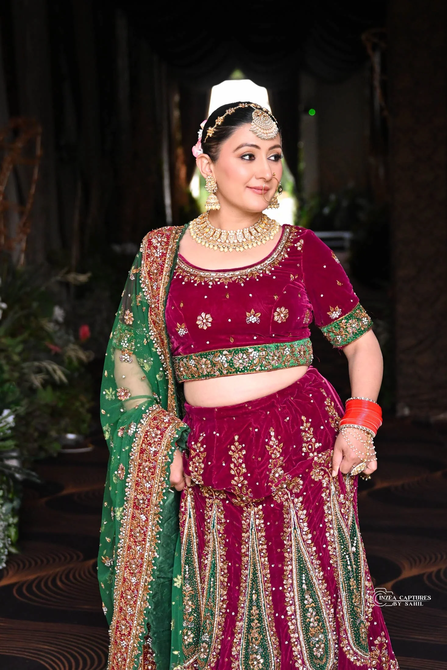A woman dressed in traditional Indian attire, wearing a red velvet lehenga with gold and green embroidery, paired with a matching blouse and a green dupatta with floral designs. She is accessorized with gold jewelry, including earrings, necklace, and