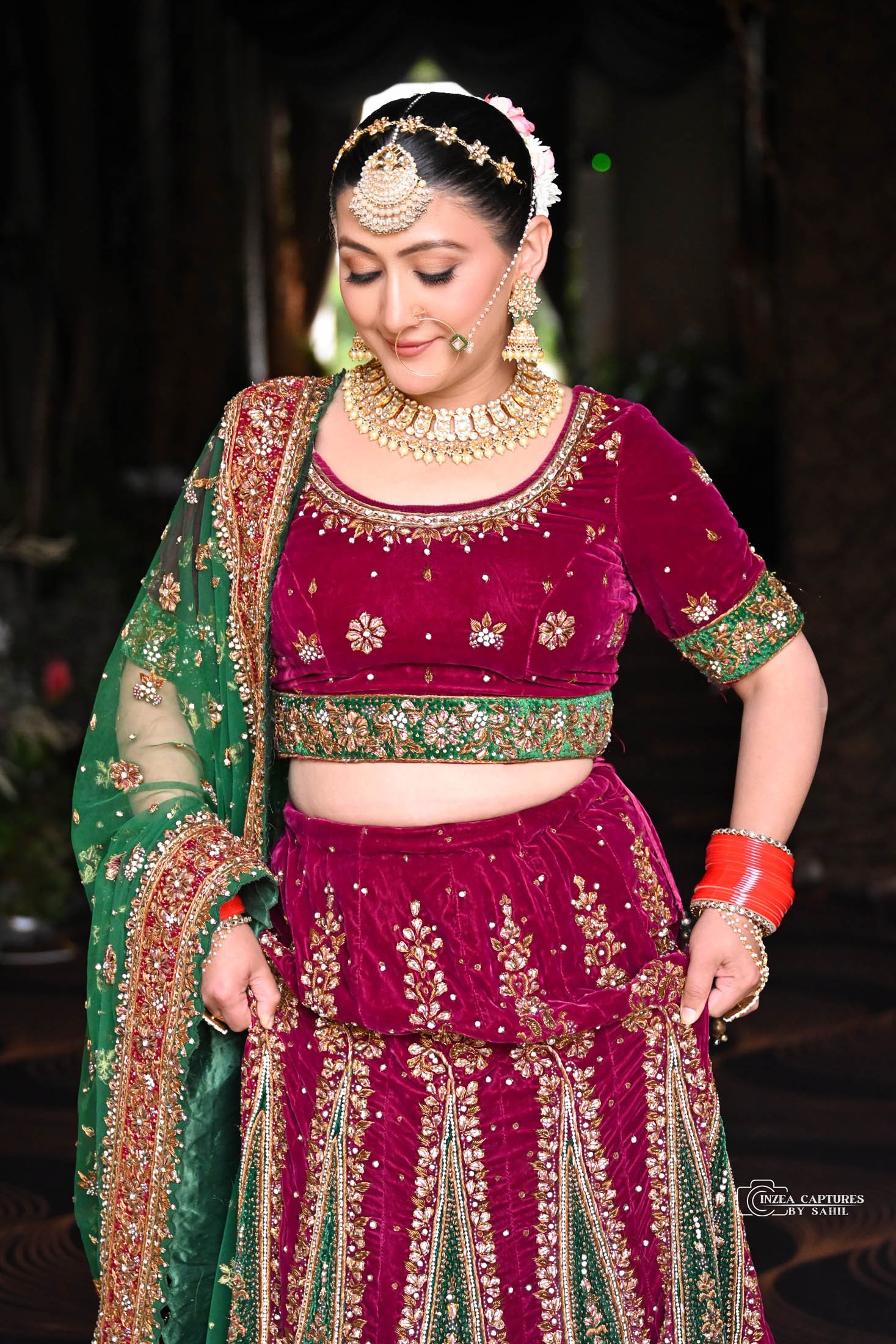 A woman dressed in traditional Indian bridal attire, wearing a maroon velvet blouse and skirt with gold embroidery, accessorized with gold jewelry and red bangles, standing in a dark setting.