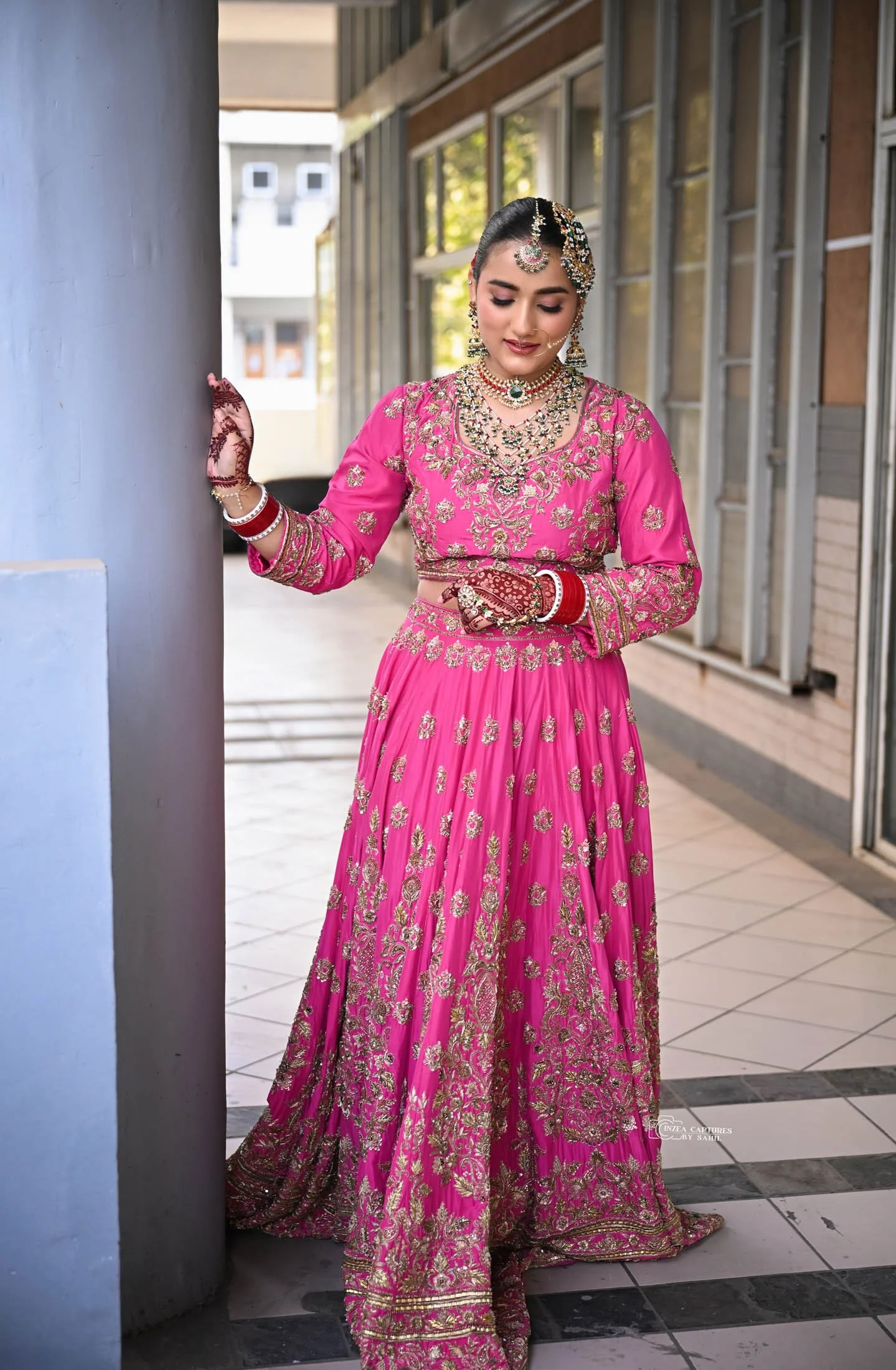 A woman dressed in a vibrant pink traditional Indian outfit with gold embroidery, adorned with jewelry including necklaces, earrings, and a headpiece, standing outdoors near a pillar.