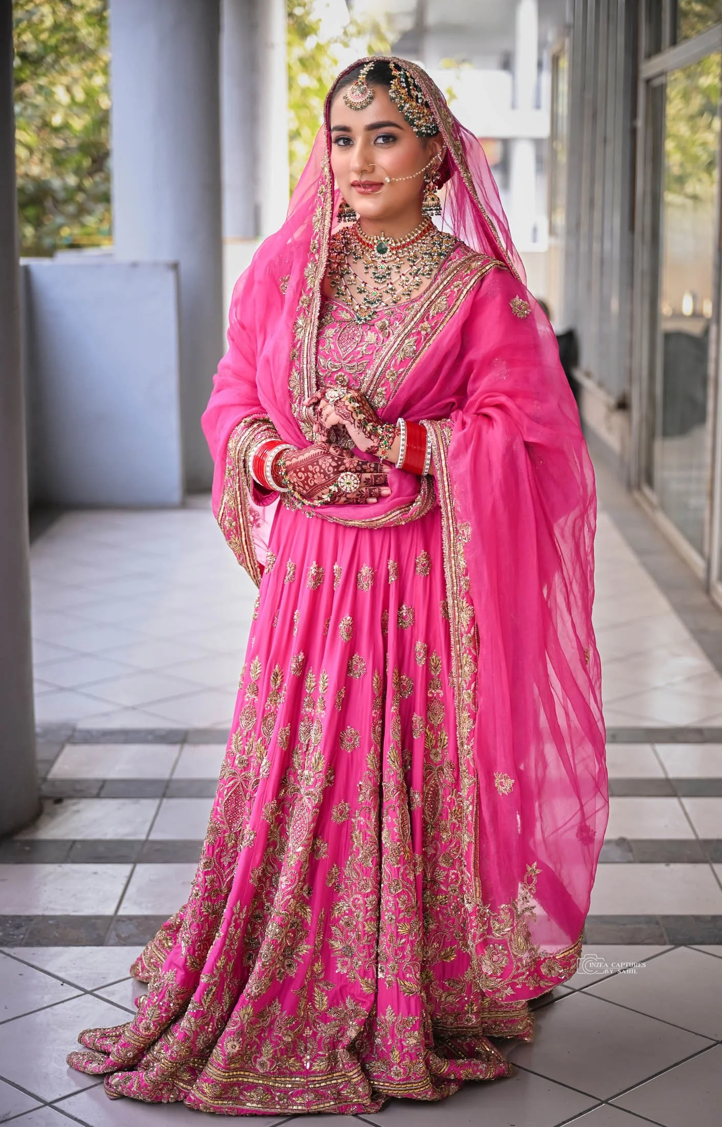 A woman dressed in traditional Indian bridal attire, wearing a vibrant pink lehenga with gold embroidery, jewelry, and a veil, standing on a tiled floor.