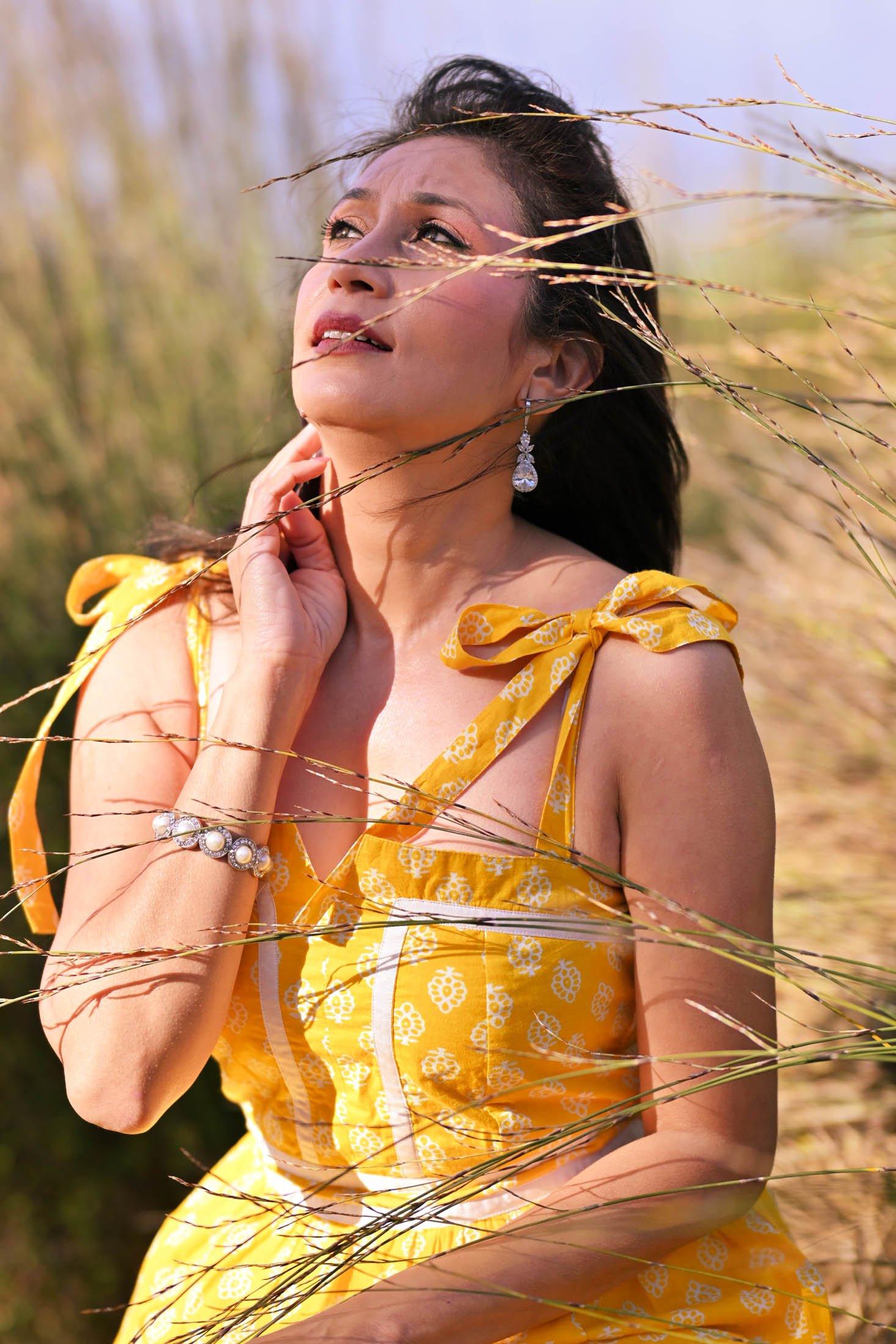 Woman in a yellow dress with floral pattern, outdoors among tall grass, wearing jewelry, gazing upward with hand on neck.