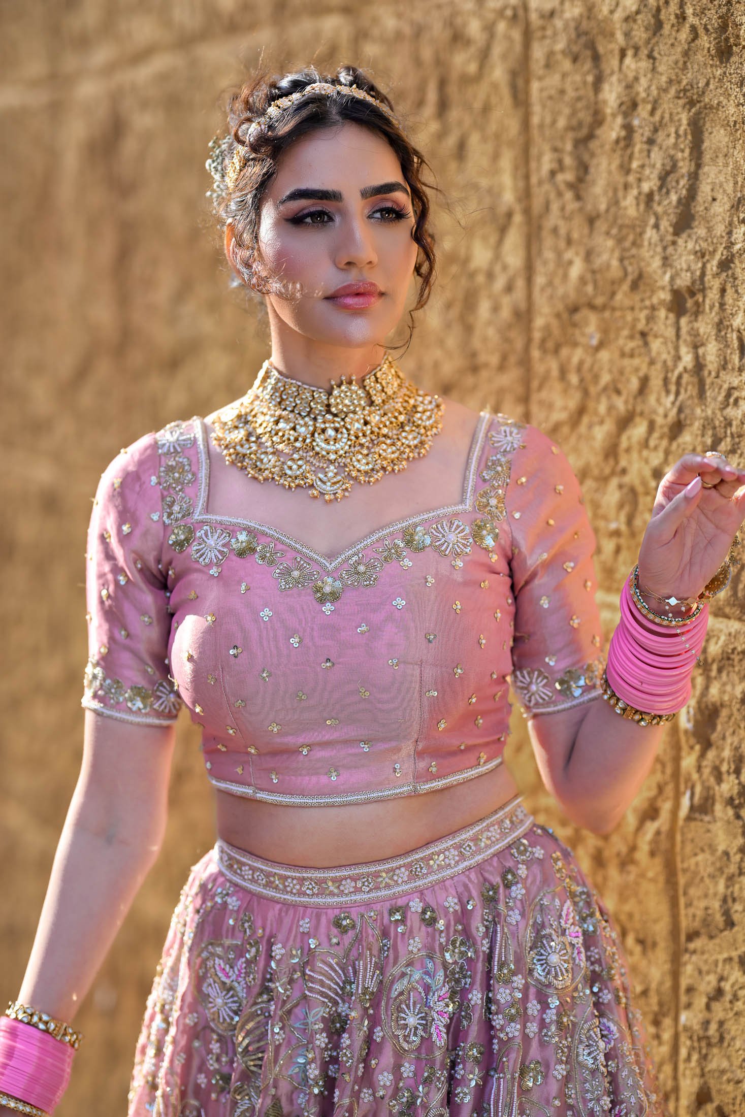 A woman wearing traditional Indian pink embroidered saree with gold embellishments, gold jewelry, and pink bangles, standing against a textured yellow wall.