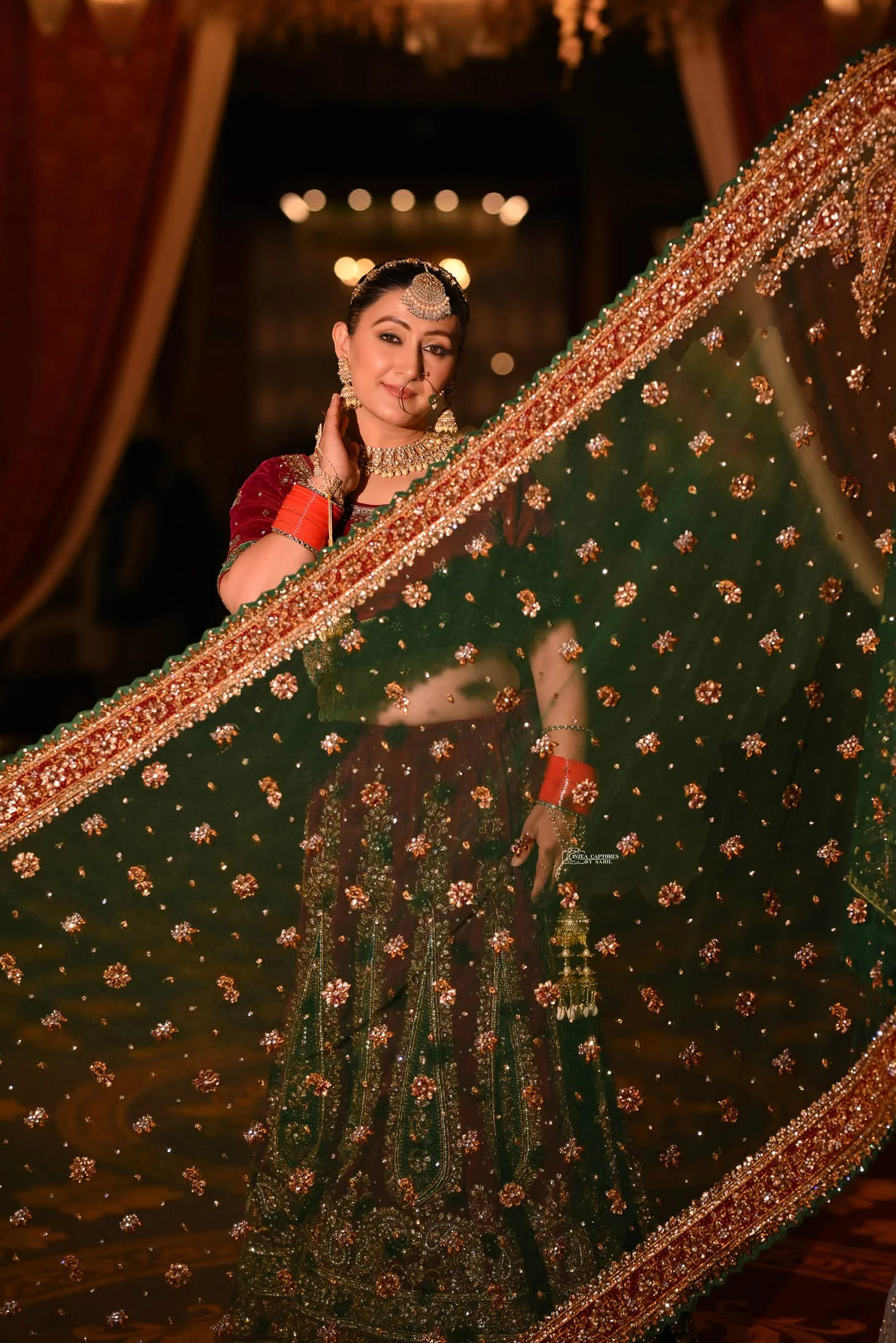 A woman in traditional Indian attire is reflected in an ornate mirror. She wears a richly embroidered green and maroon lehenga, adorned with gold jewelry and red bangles. She has a neutral-colored nose ring, large earrings, and a headpiece. The backg