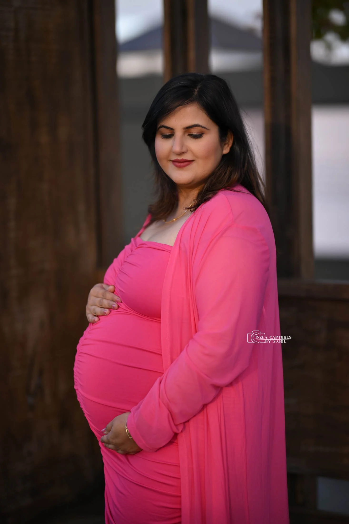 Pregnant woman in pink dress gently holding her belly and smiling with closed eyes outdoors.