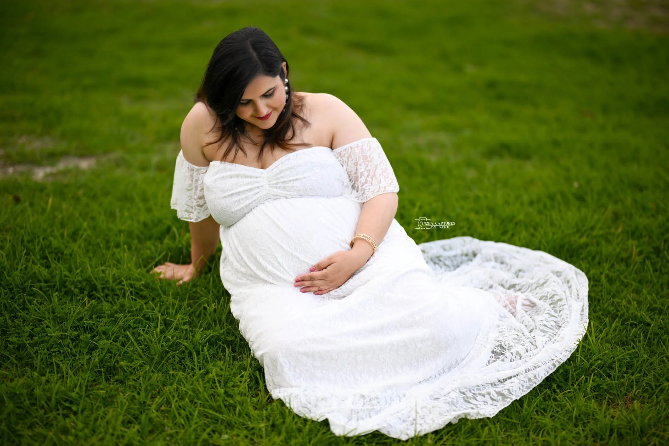 A pregnant woman sitting on green grass, wearing a white lace off-shoulder dress, with her hand on her belly, looking down and smiling gently.
