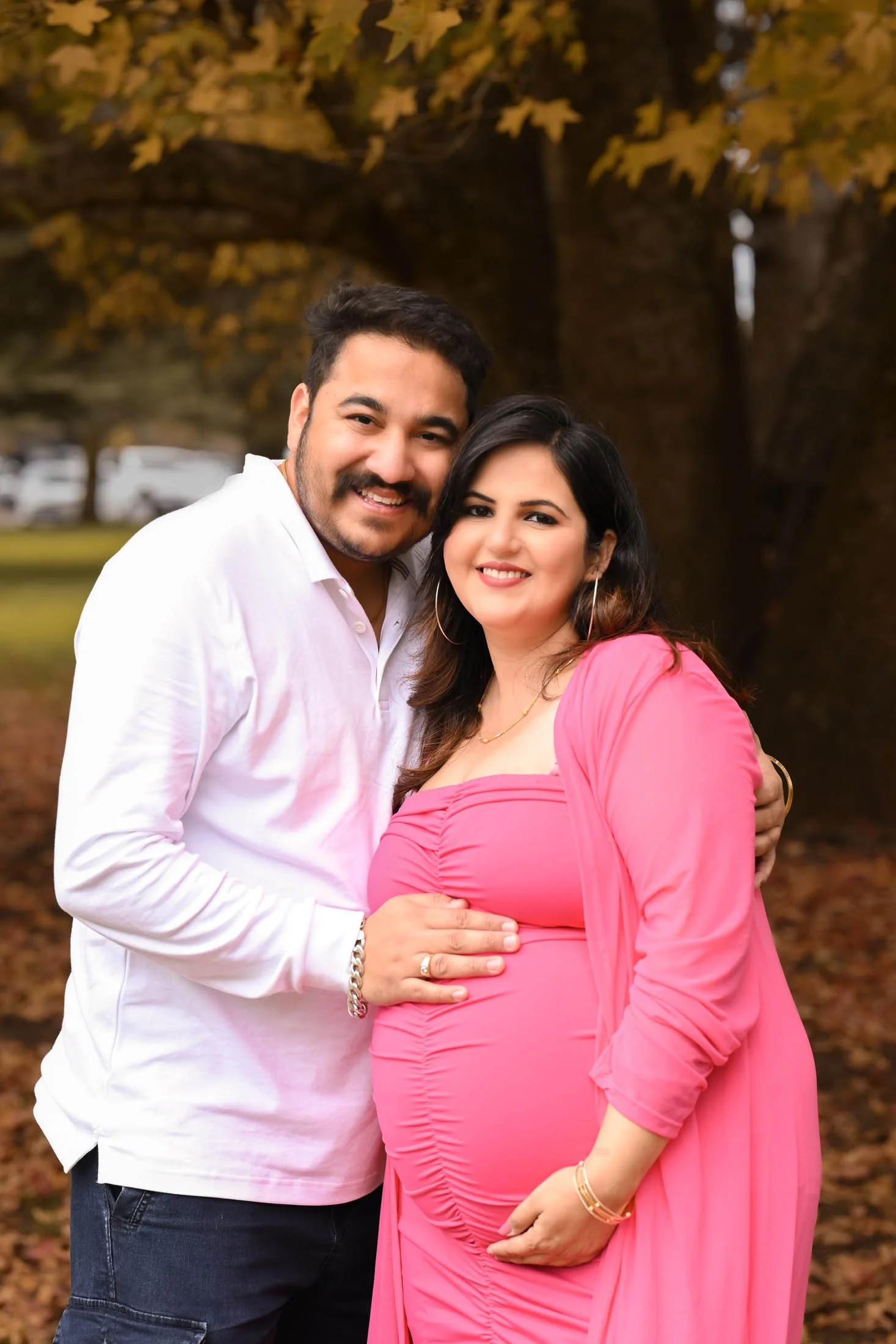 A happy couple outdoors, the man wearing a white shirt and the pregnant woman in a pink dress, embracing each other in front of a large tree with autumn leaves.