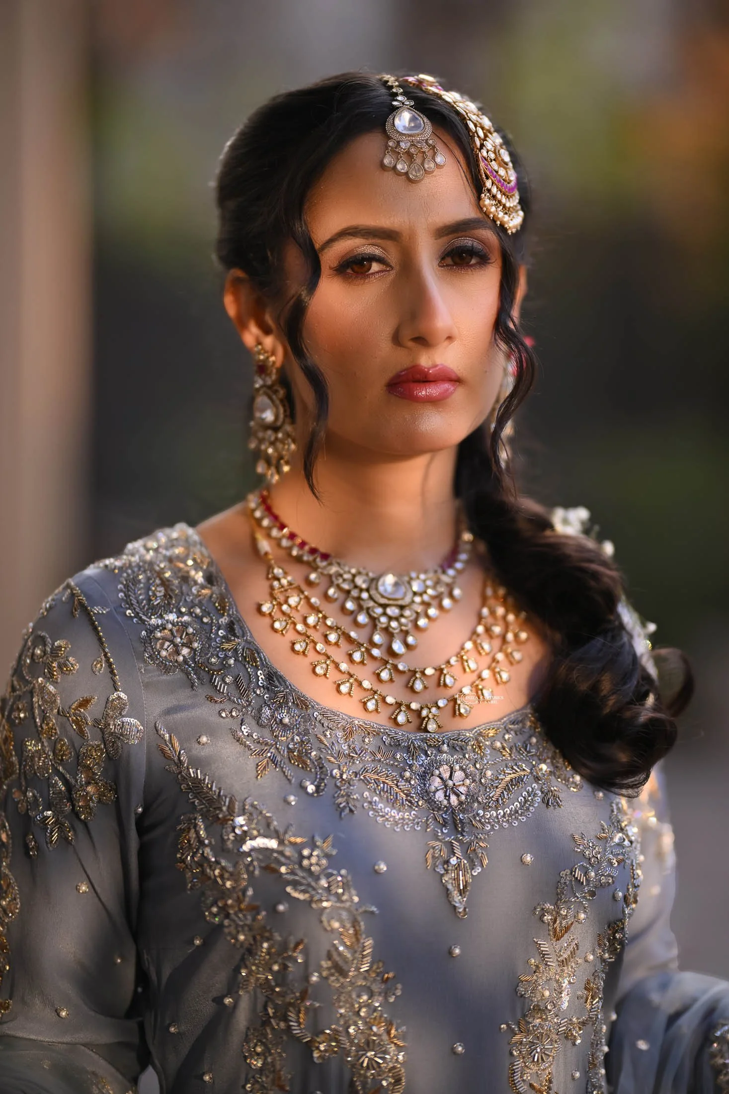 A woman dressed in traditional Indian attire with elaborate jewelry, including necklaces, earrings, and a headpiece, posing outdoors with a blurred background.