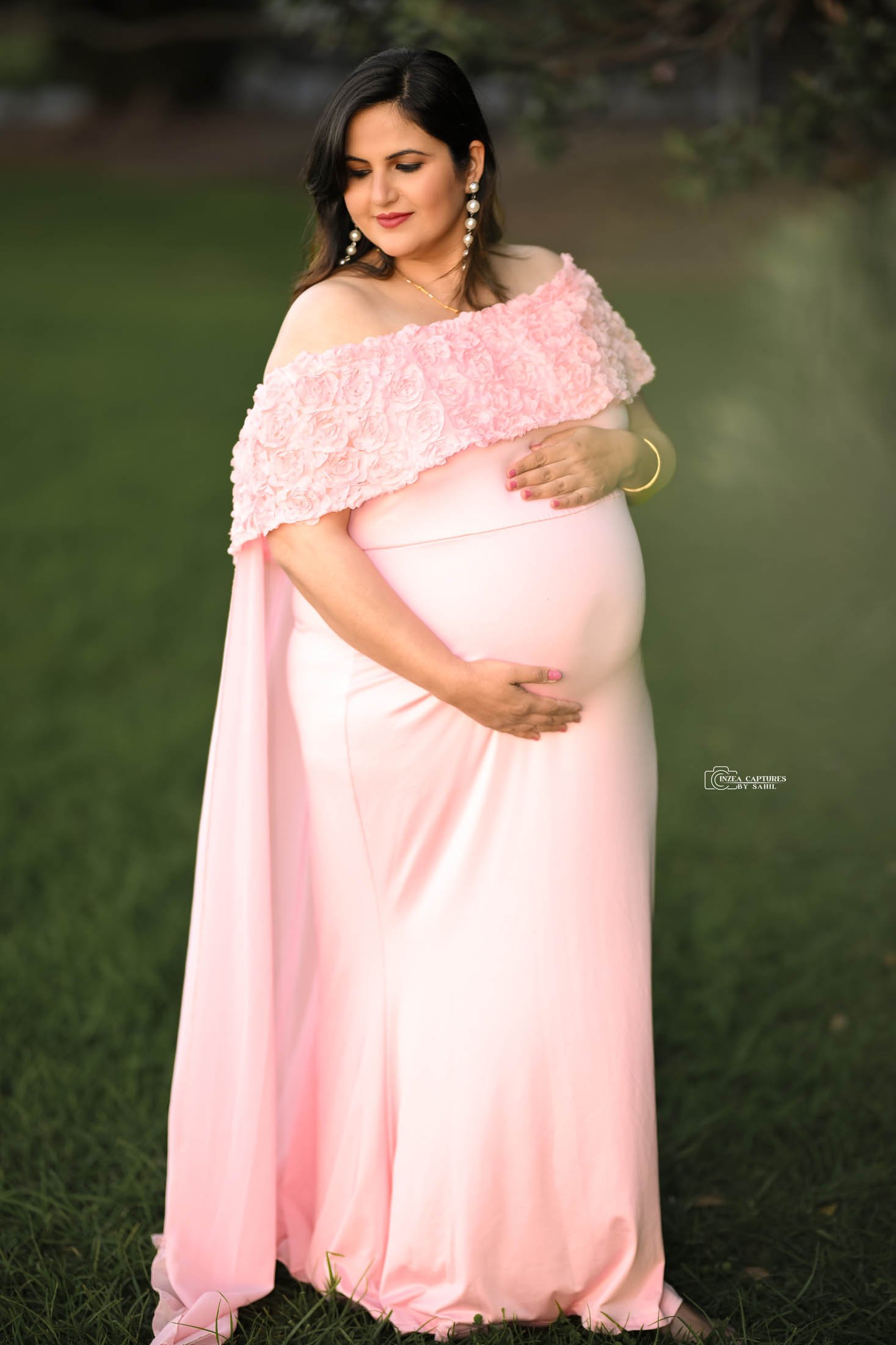 A pregnant woman in a pink dress with floral details on the top, standing outdoors on grass, gently holding her belly with one hand, and her other hand resting on her chest. She is wearing earrings, a necklace, and a bracelet.